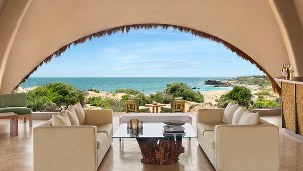 Una habitación con mesa de vidrio y dos sofás orientados hacia una vista a la playa a través de un ventanal arqueado.