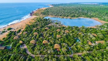 Une vue aérienne d’un complexe hôtelier avec des bungalows aux toits de chaume, entourés d’une végétation luxuriante, d’une plage et d’une vaste étendue d’eau.