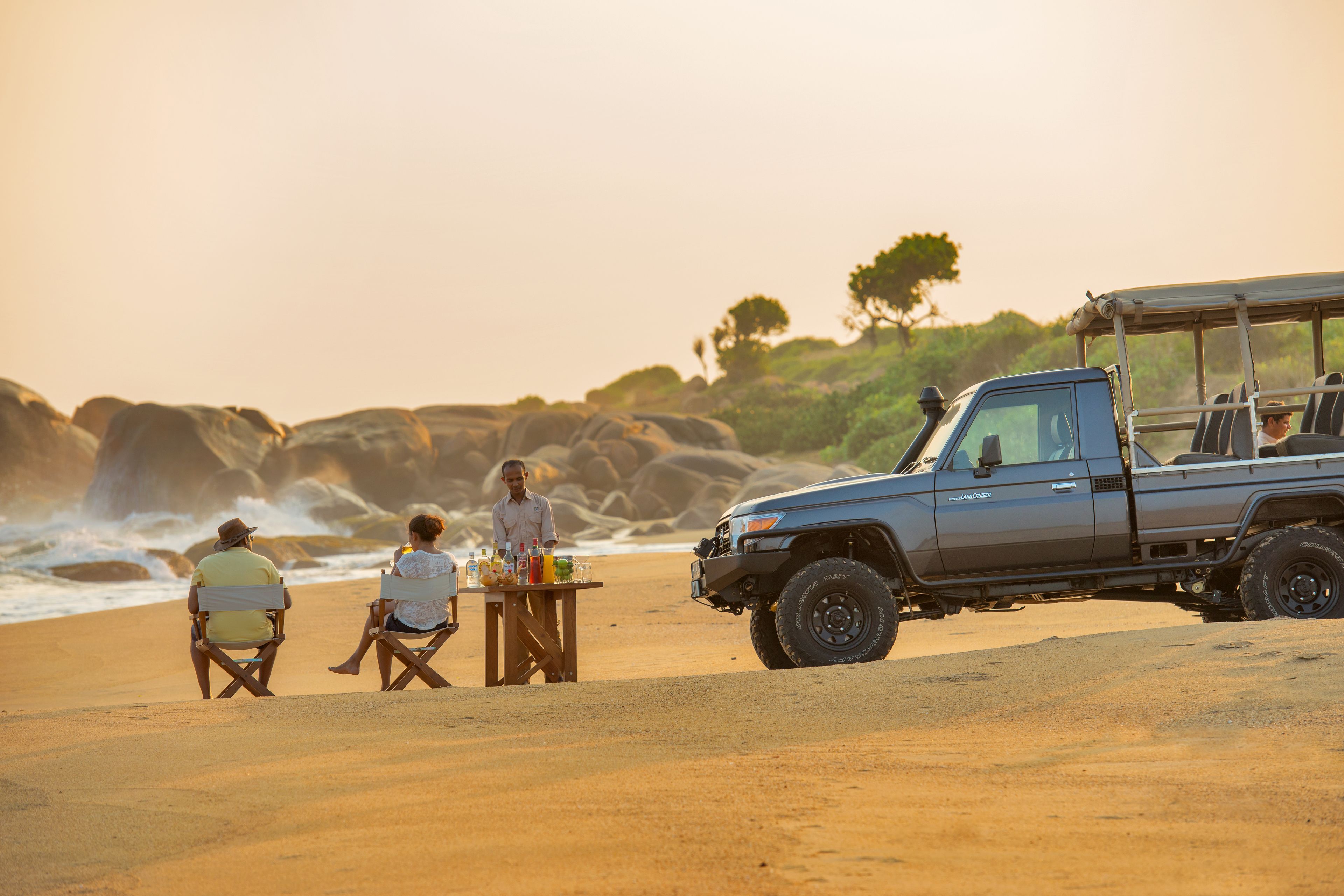 A beach scene with a vehicle and two people sitting on chairs.