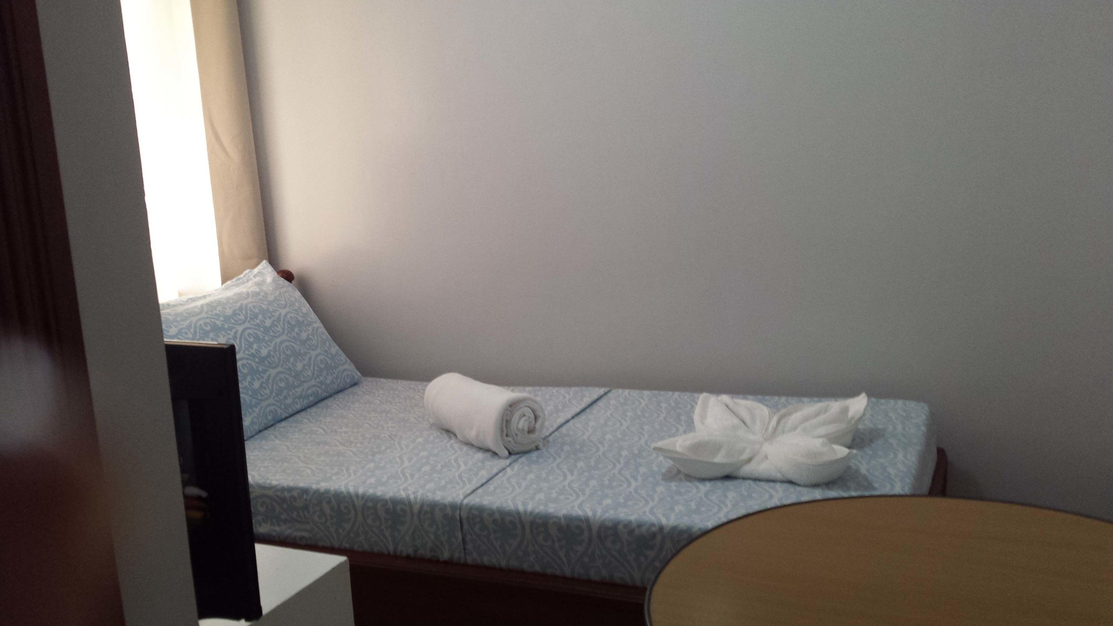 A small, single-bed room with a light blue patterned bedspread, a rolled white towel, and a white pillow.