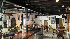 Angolo bar con un bancone in legno, sgabelli da bar e un rubinetto per la birra. Sullo sfondo, tavoli e sedie, e un ventilatore a parete.