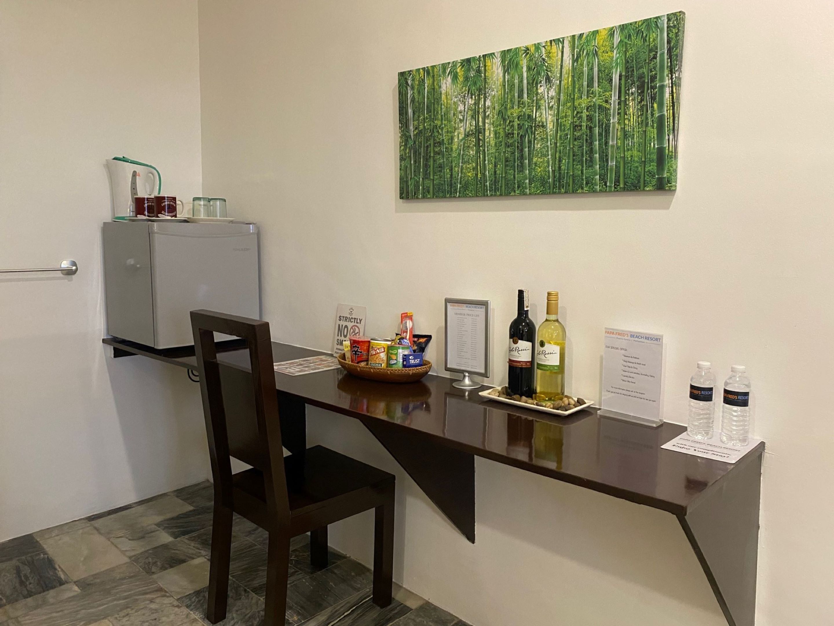 A small office with a desk, chair, and a refrigerator. There is a painting of a forest on the wall, a menu sign, and a bottle of wine.
