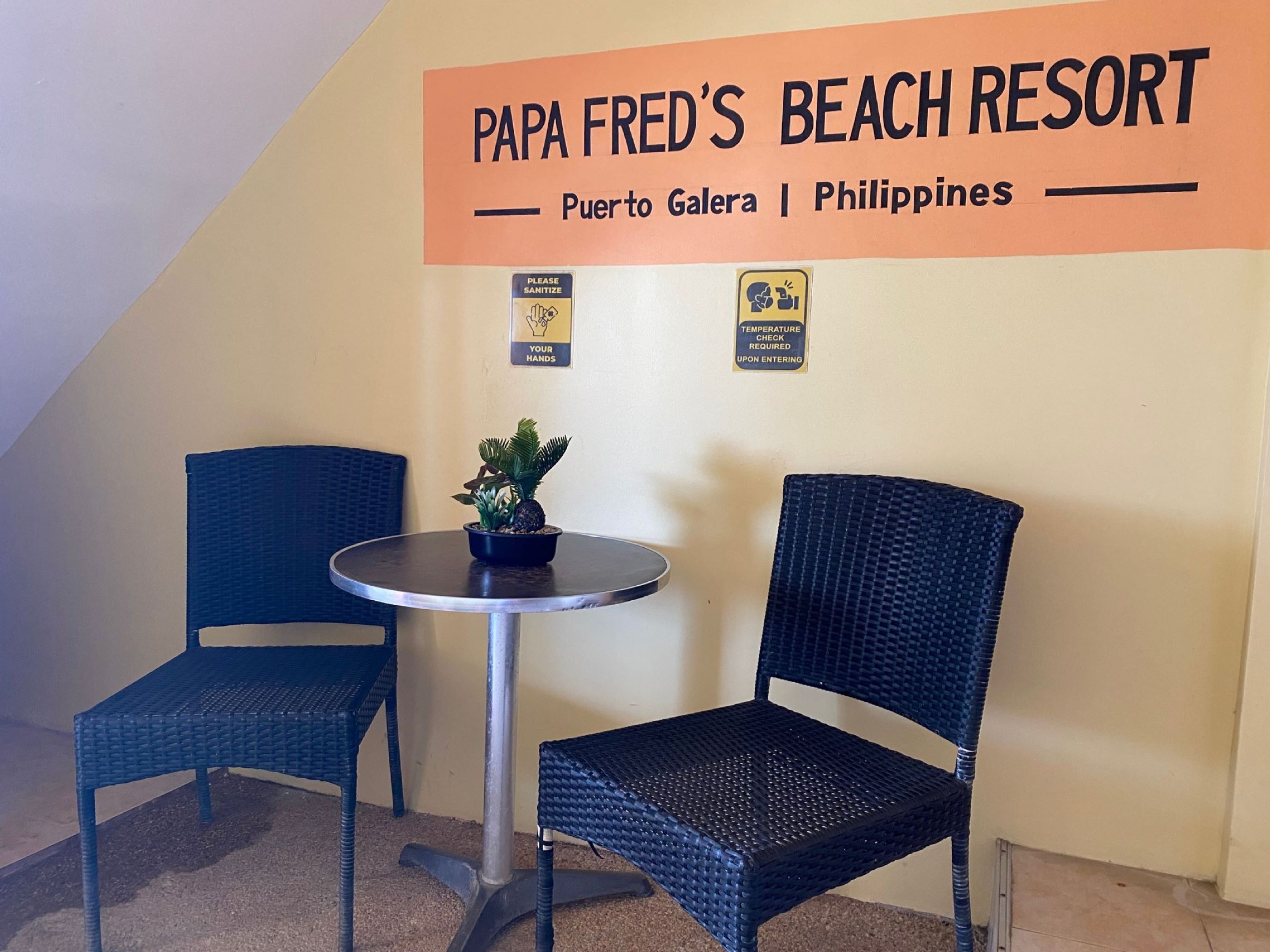 A small seating area with two wicker chairs and a round table with a potted plant. A sign above reads "Papa Fred's Beach Resort, Puerto Galera, Philippines." Two health and safety signs are also visible.