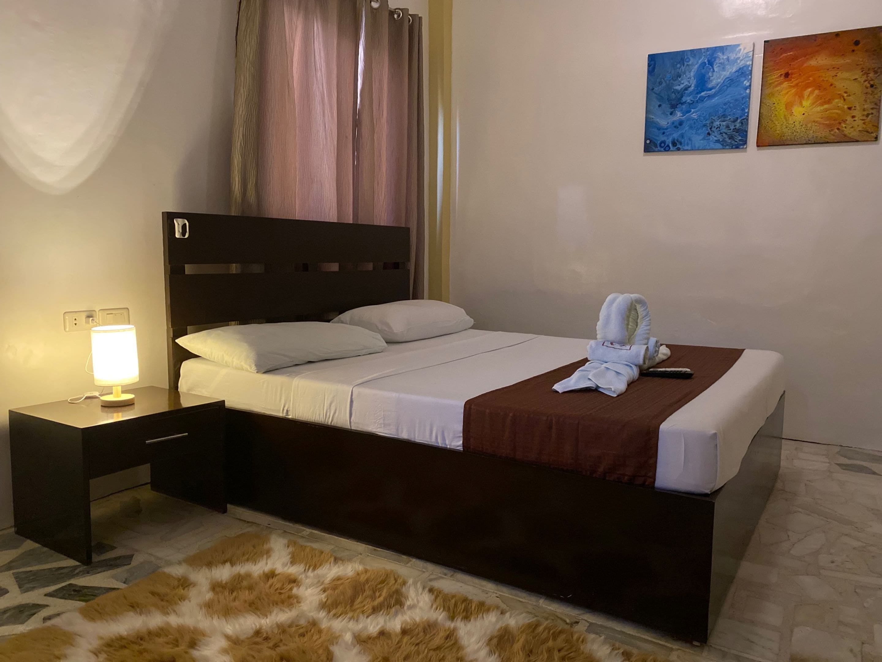 Una cama bien hecha con ropa blanca, un cabecero marrón y un control remoto sobre la cama. Hay una alfombra mullida en el suelo y una mesita de noche con una lámpara.