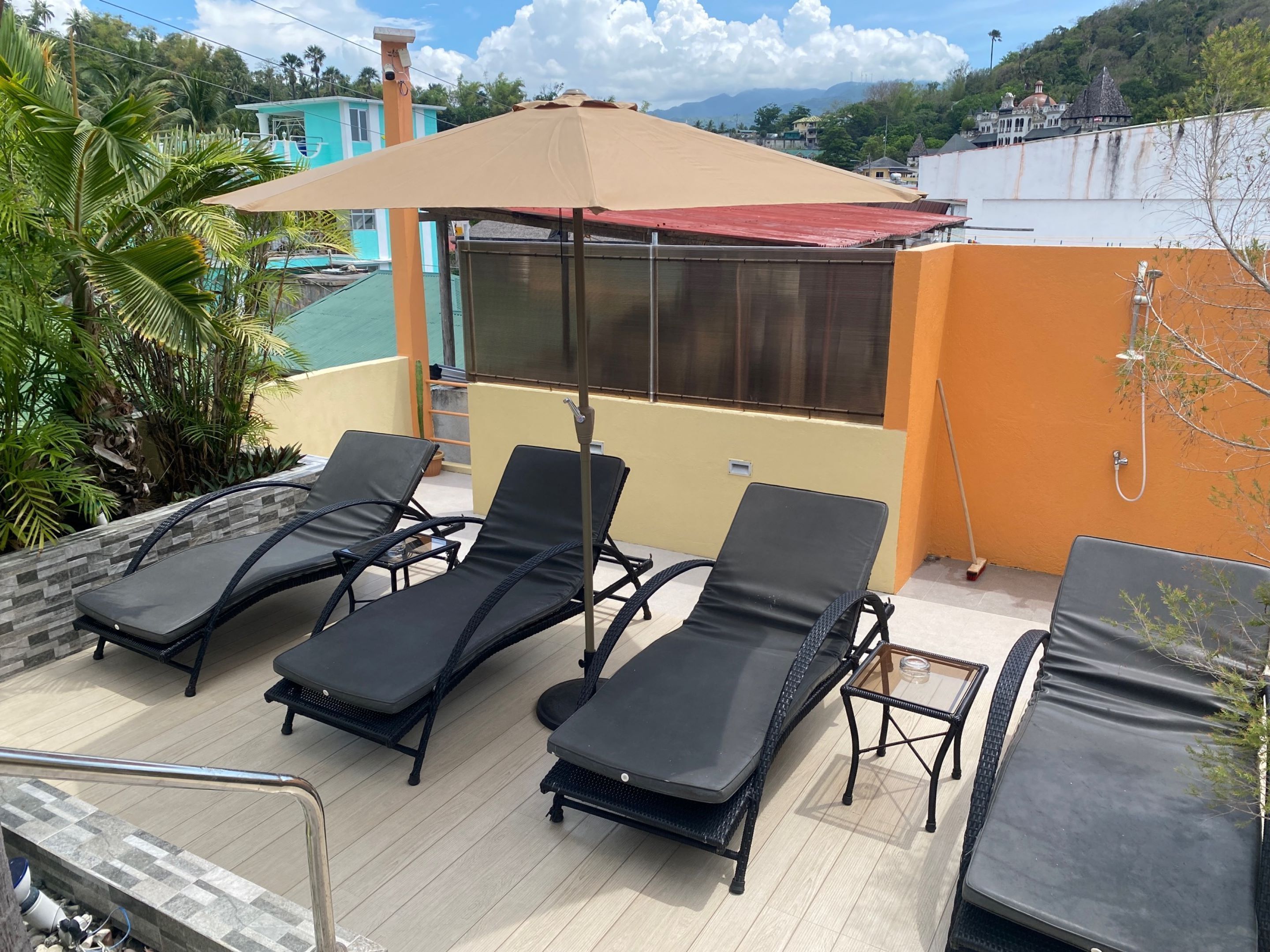 A rooftop terrace with four lounge chairs, a large umbrella, and a view of a pool and buildings in the background.
