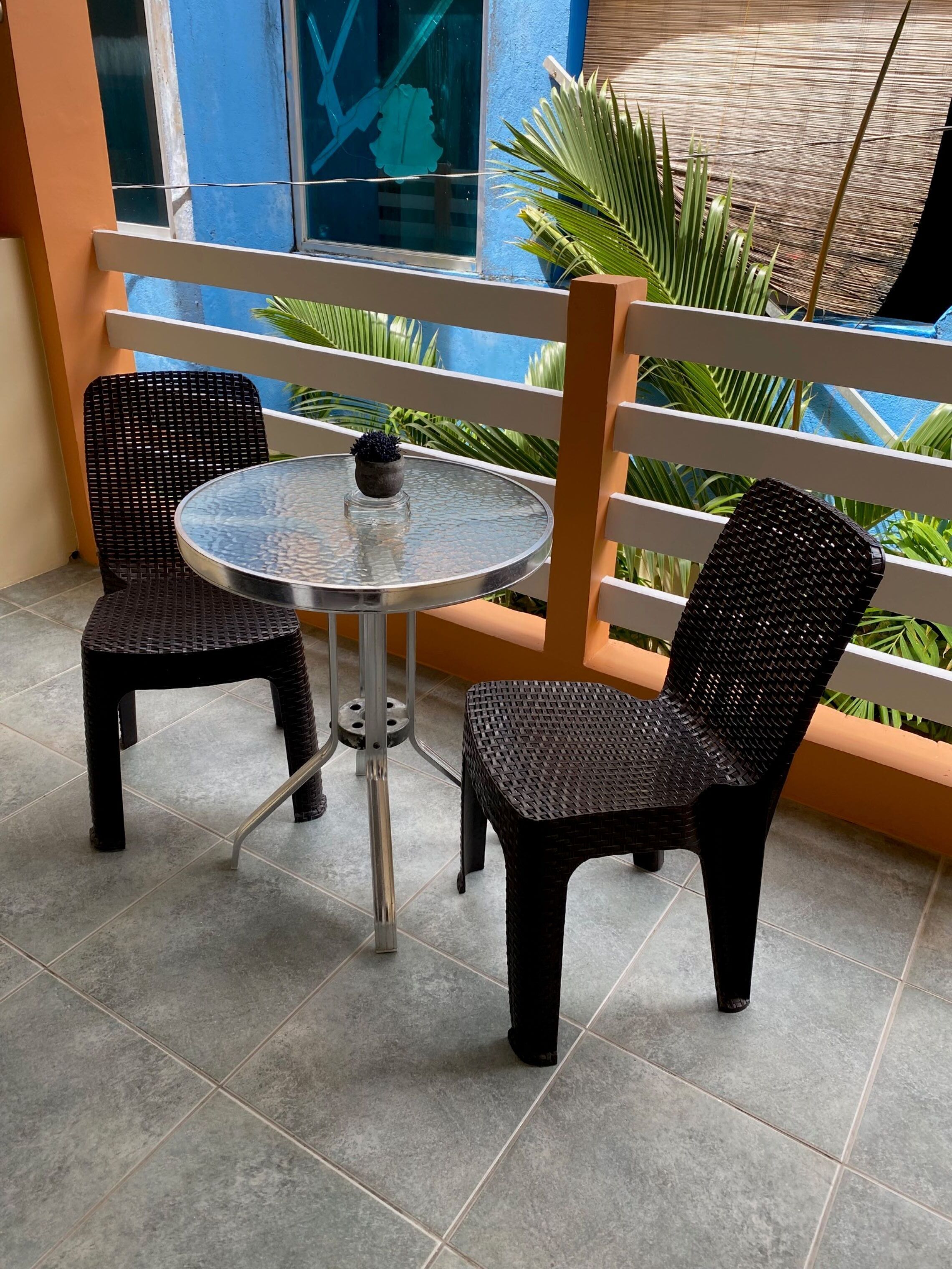 Ein Balkon mit zwei Korbstühlen und einem kleinen Tisch, umgeben von einem Geländer und Blumentöpfen.