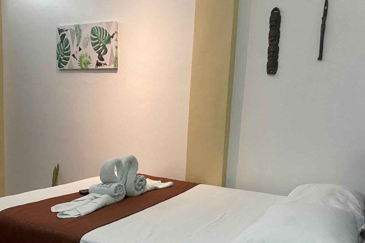 A neatly made bed with white linens and a brown throw blanket, a small plant beside the bed, and a wall-mounted artwork of tropical leaves.
