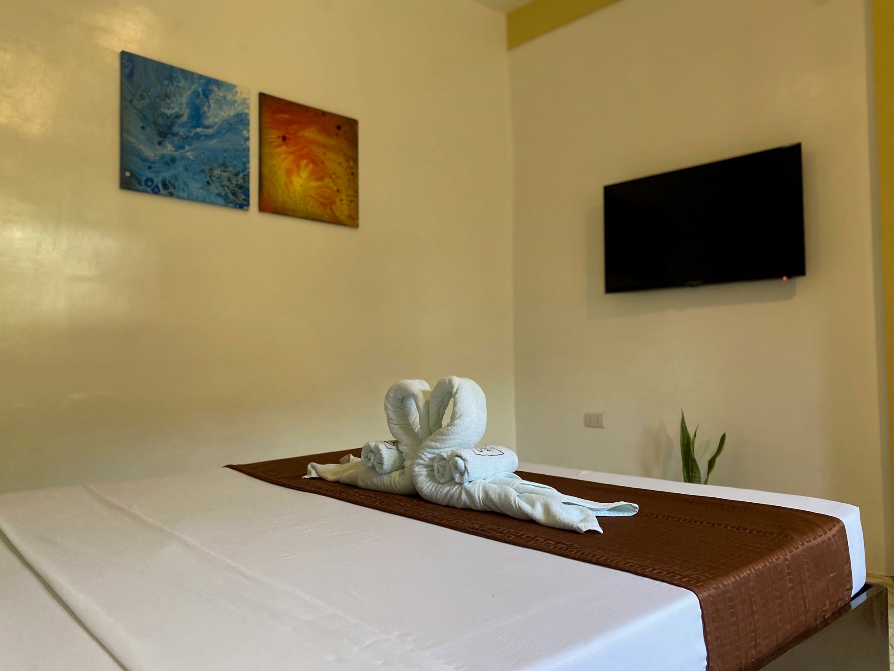 A neatly made bed with a brown headboard, a white bedspread, and a folded towel on the bed. There are two paintings on the wall above the bed and a flat-screen TV mounted on the wall.