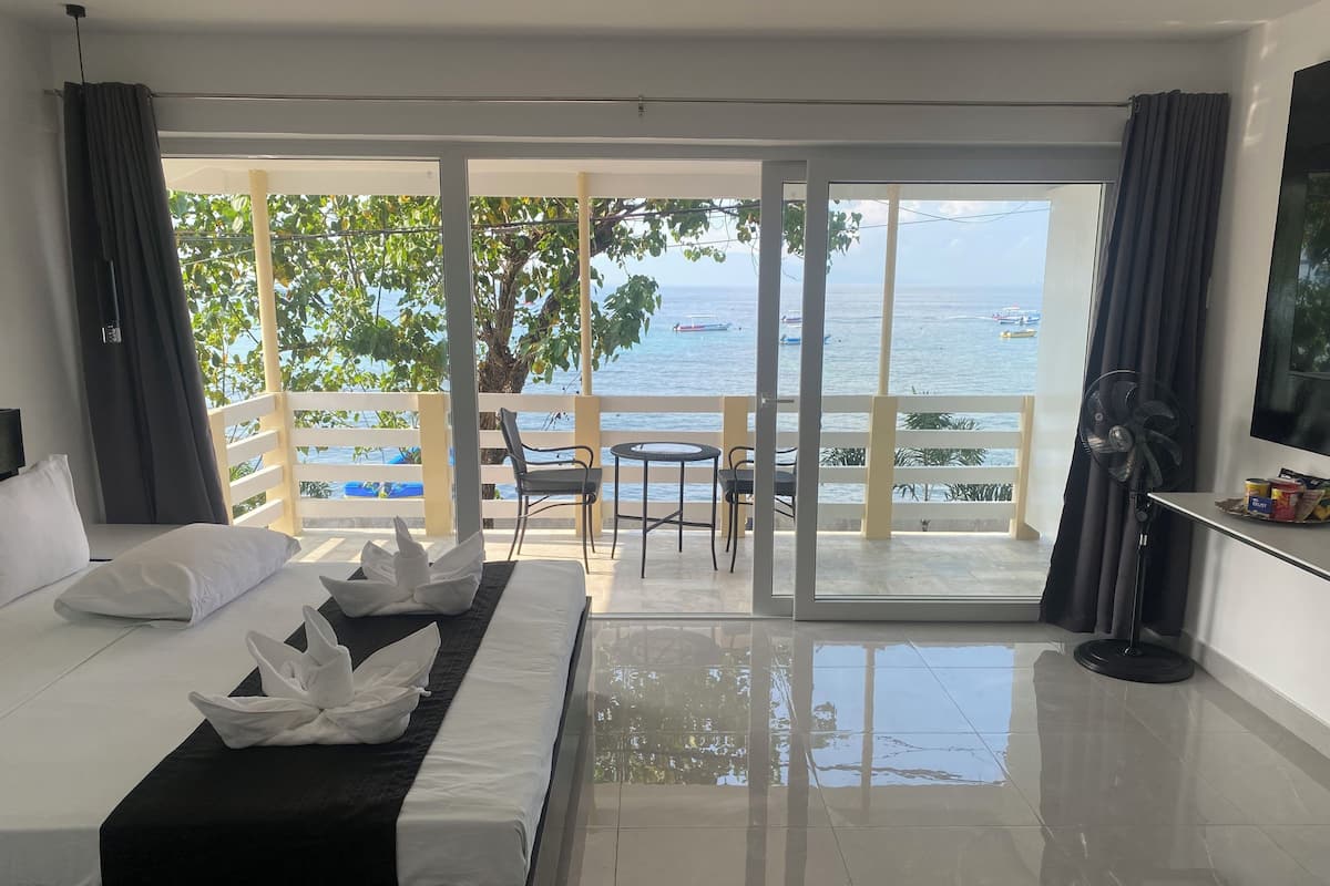 A hotel room with a bed, a small table, a chair, and a balcony with a view of the sea.