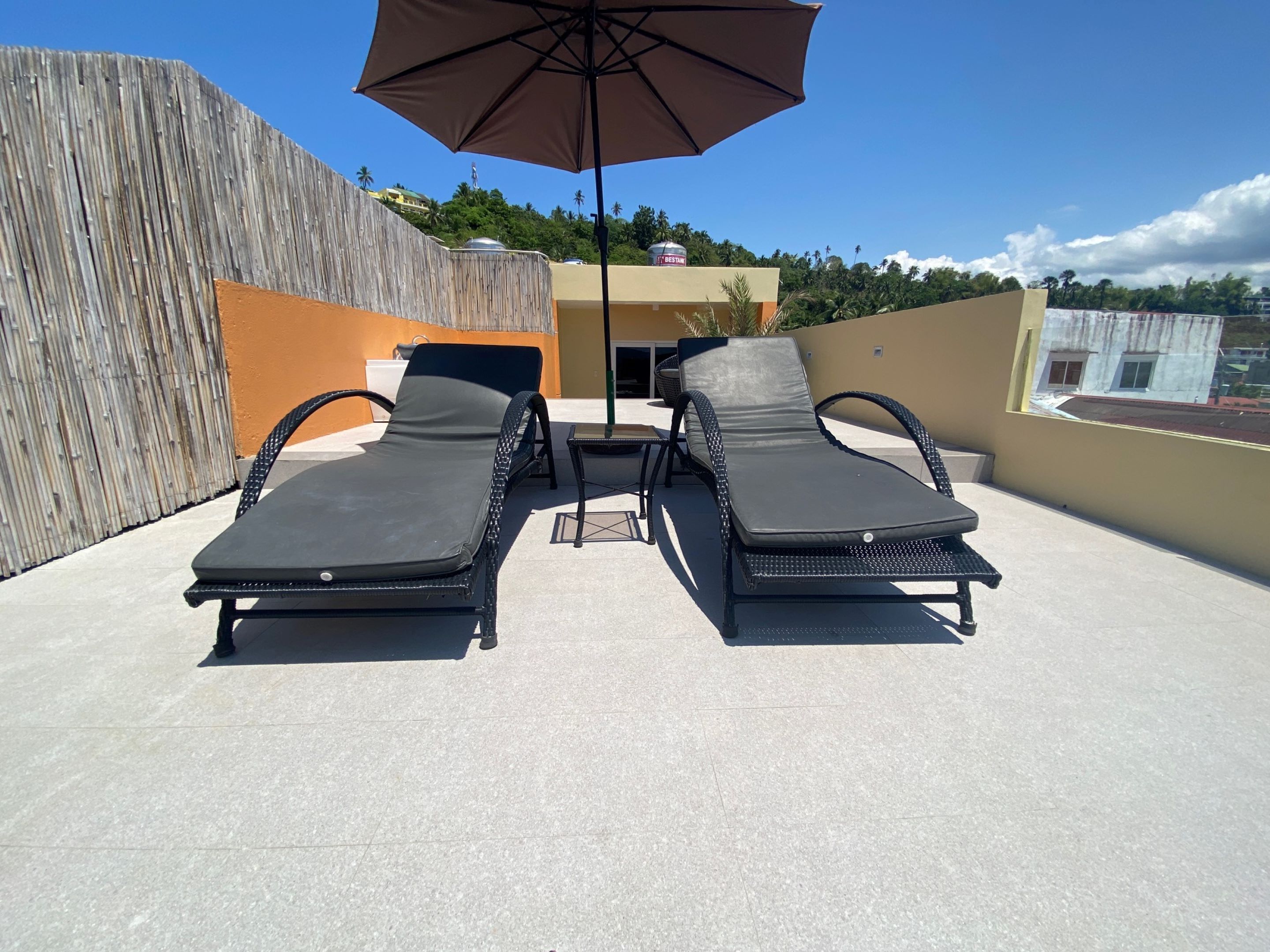 A rooftop terrace with two lounge chairs and a table, a large umbrella, and a view of a building with a sign that reads "BESTAM".