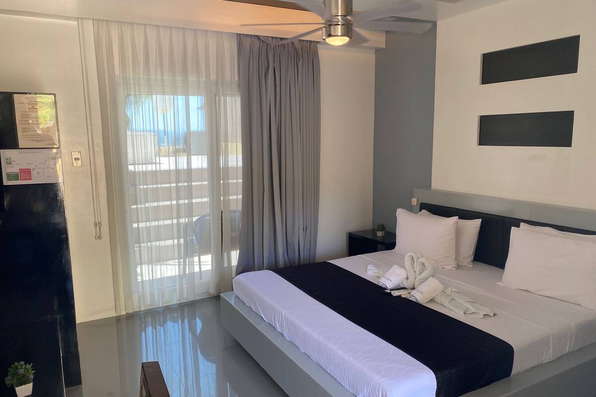 A modern hotel room with a large bed, a ceiling fan, a small bedside table with a plant, and a view through a large window with sheer curtains.