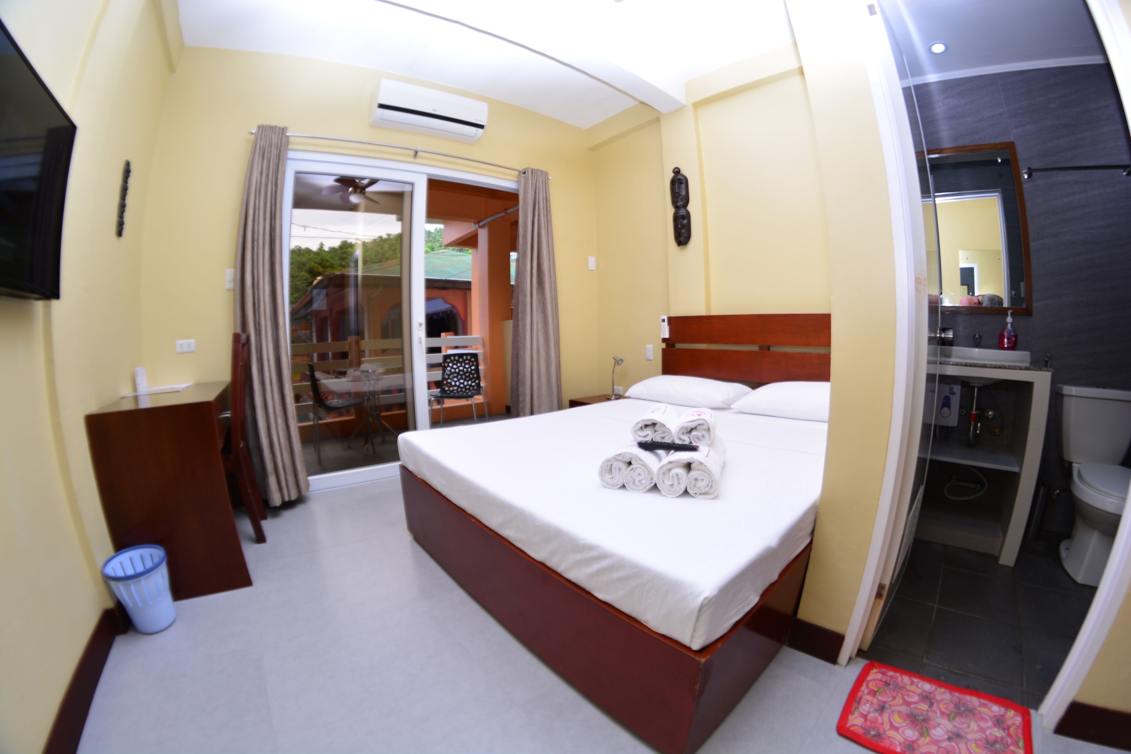 Une chambre d’hôtel avec un lit, un bureau, une chaise, une petite poubelle et une salle de bain visible à travers une porte ouverte.