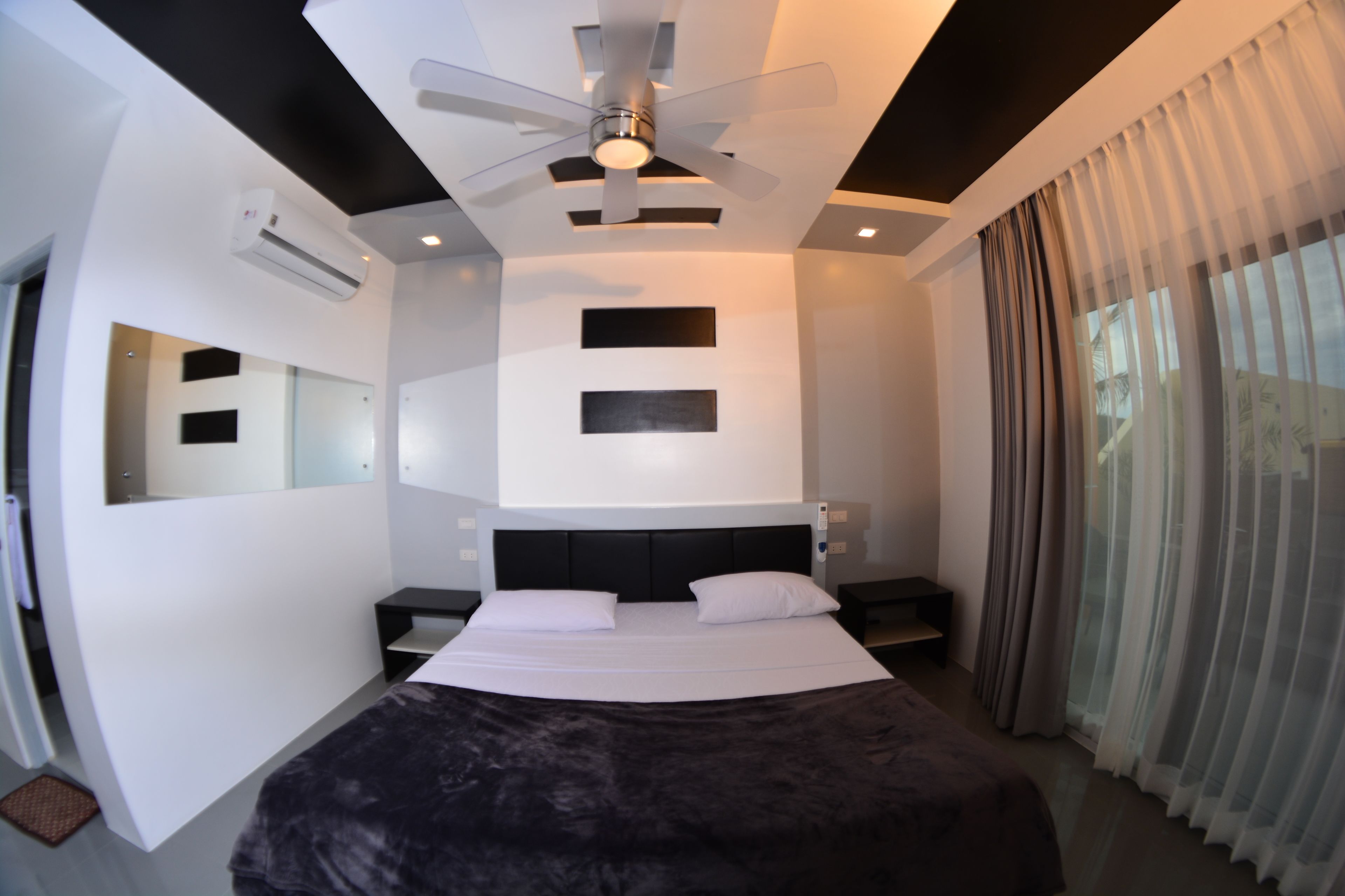 A modern hotel room with a large bed, a ceiling fan, a flat-screen TV, and a large window with curtains.
