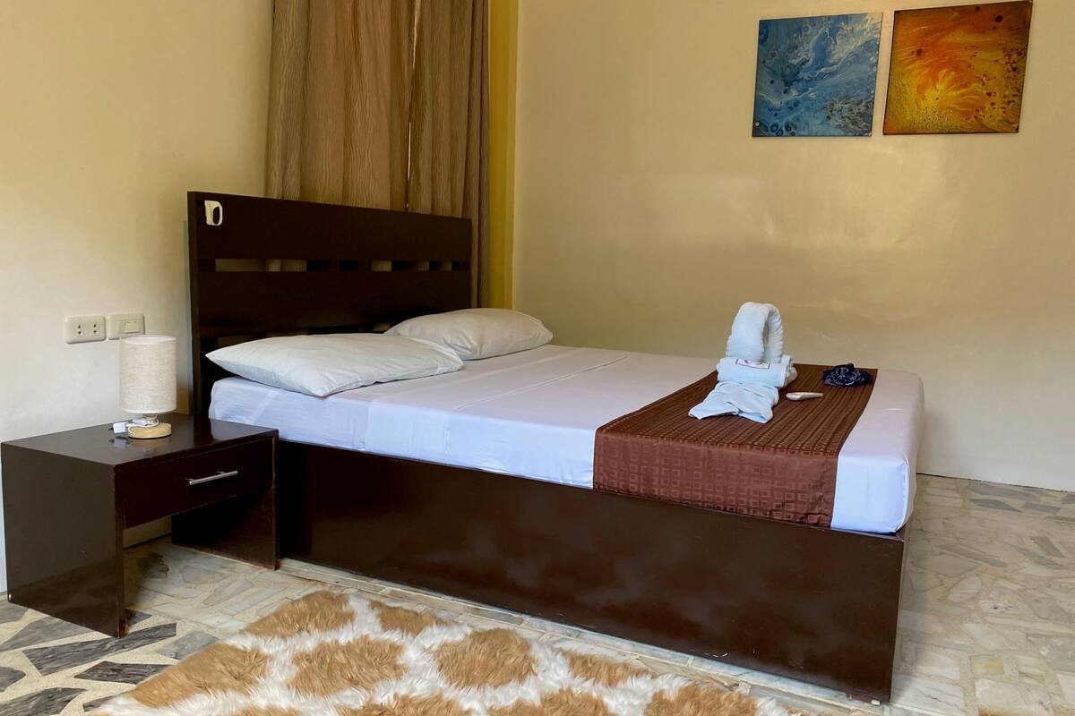 A neatly made bed with a brown headboard, a bedside table with a lamp, and a patterned rug on the floor.
