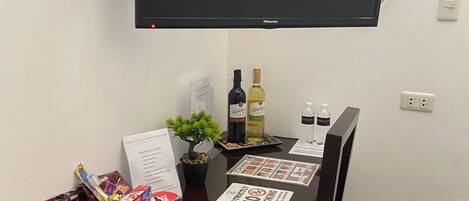 Un área de comedor con un televisor de pantalla plana montado, una mesa con un mantel individual, un menú, una botella de vino y varios aperitivos.