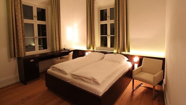 Un dormitorio con una cama doble, una mesita de noche, una silla y dos ventanas con cortinas.