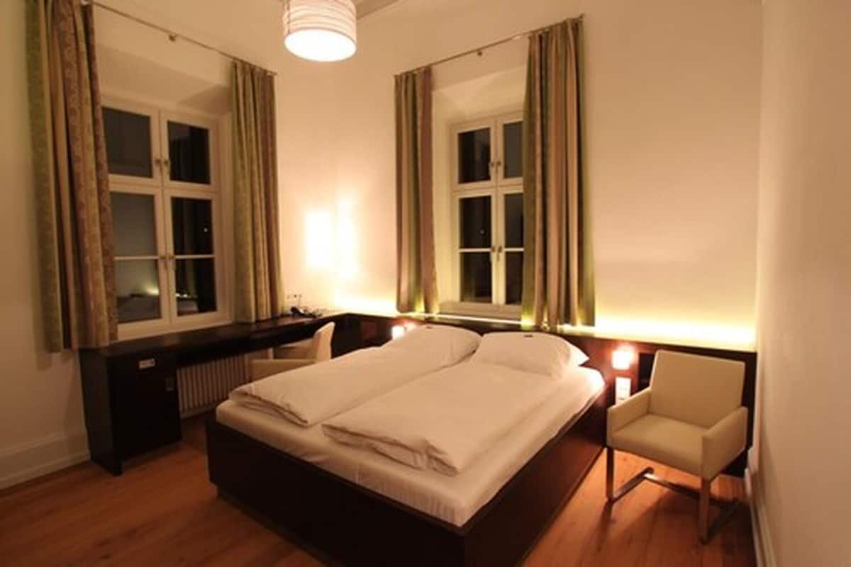 Een slaapkamer met een tweepersoonsbed, een nachtkastje, een stoel en twee ramen met gordijnen.
