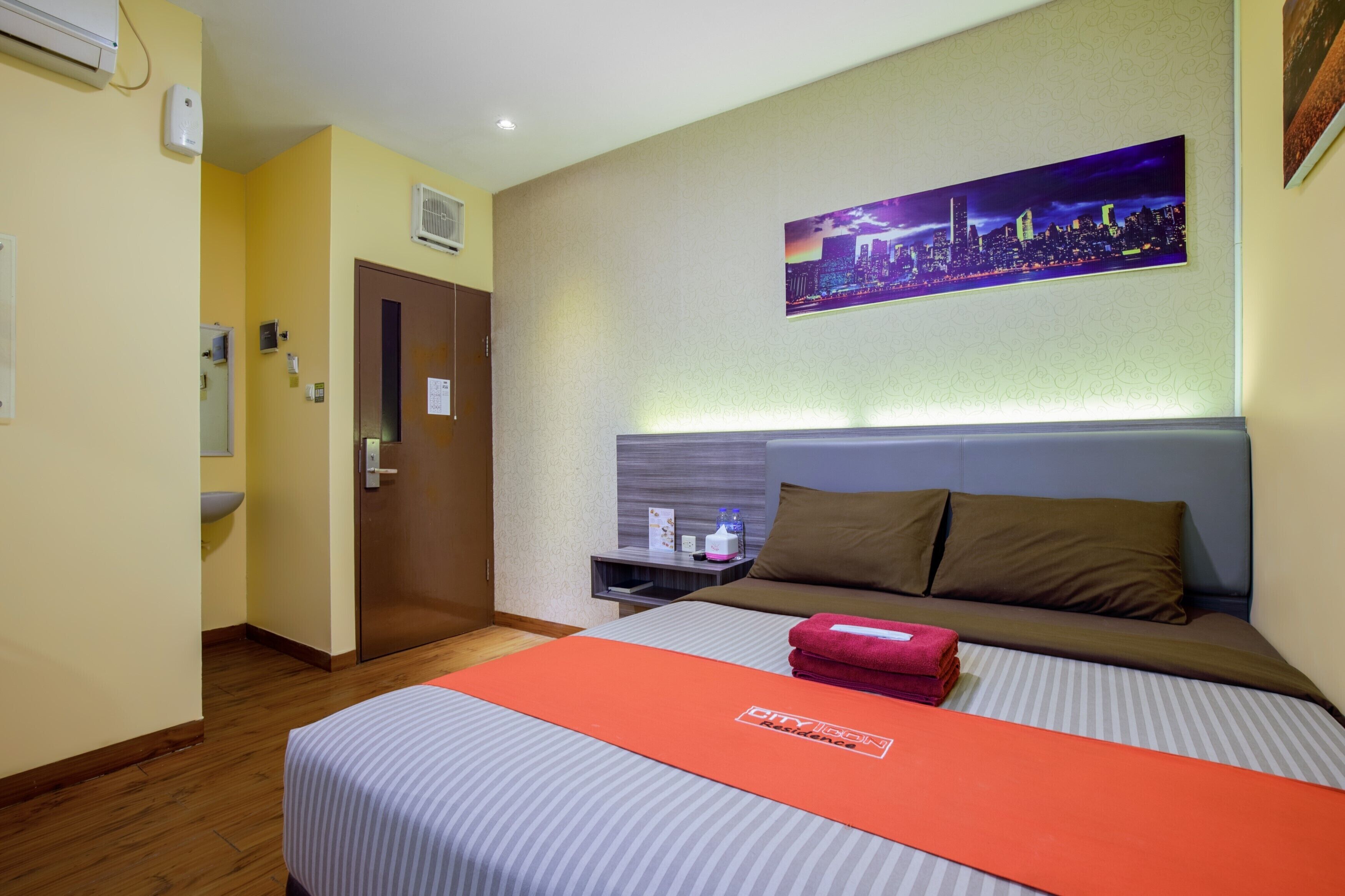 A hotel room with a bed, a nightstand, a cityscape painting, and a door.