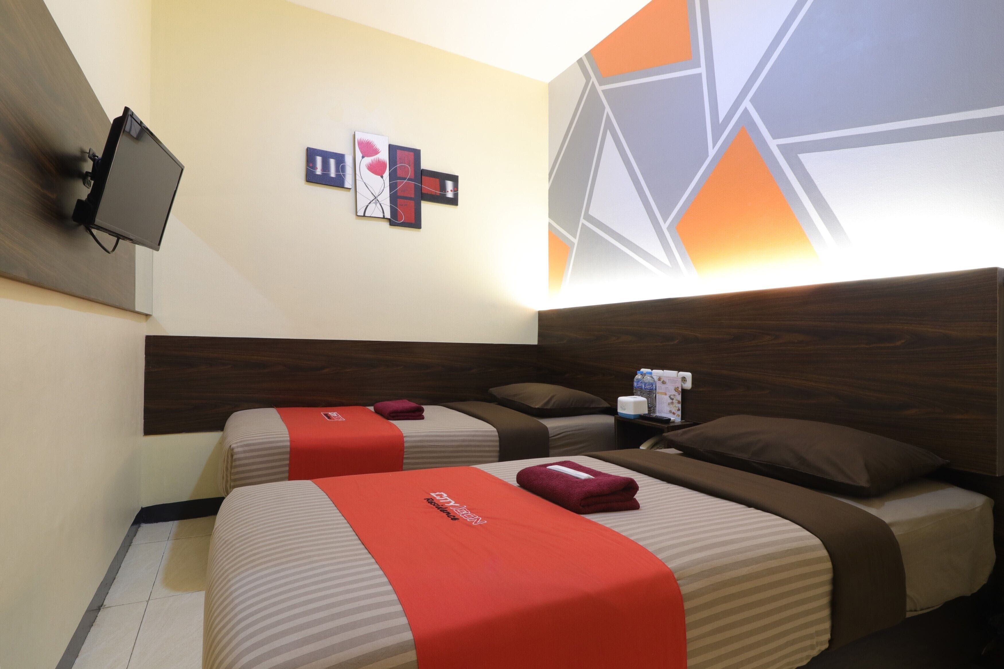 A hotel room with two beds, each with a red bedspread and a pillow. There is a small table with a bottle and a tissue box. The room has a wooden headboard and a wall with geometric patterns.