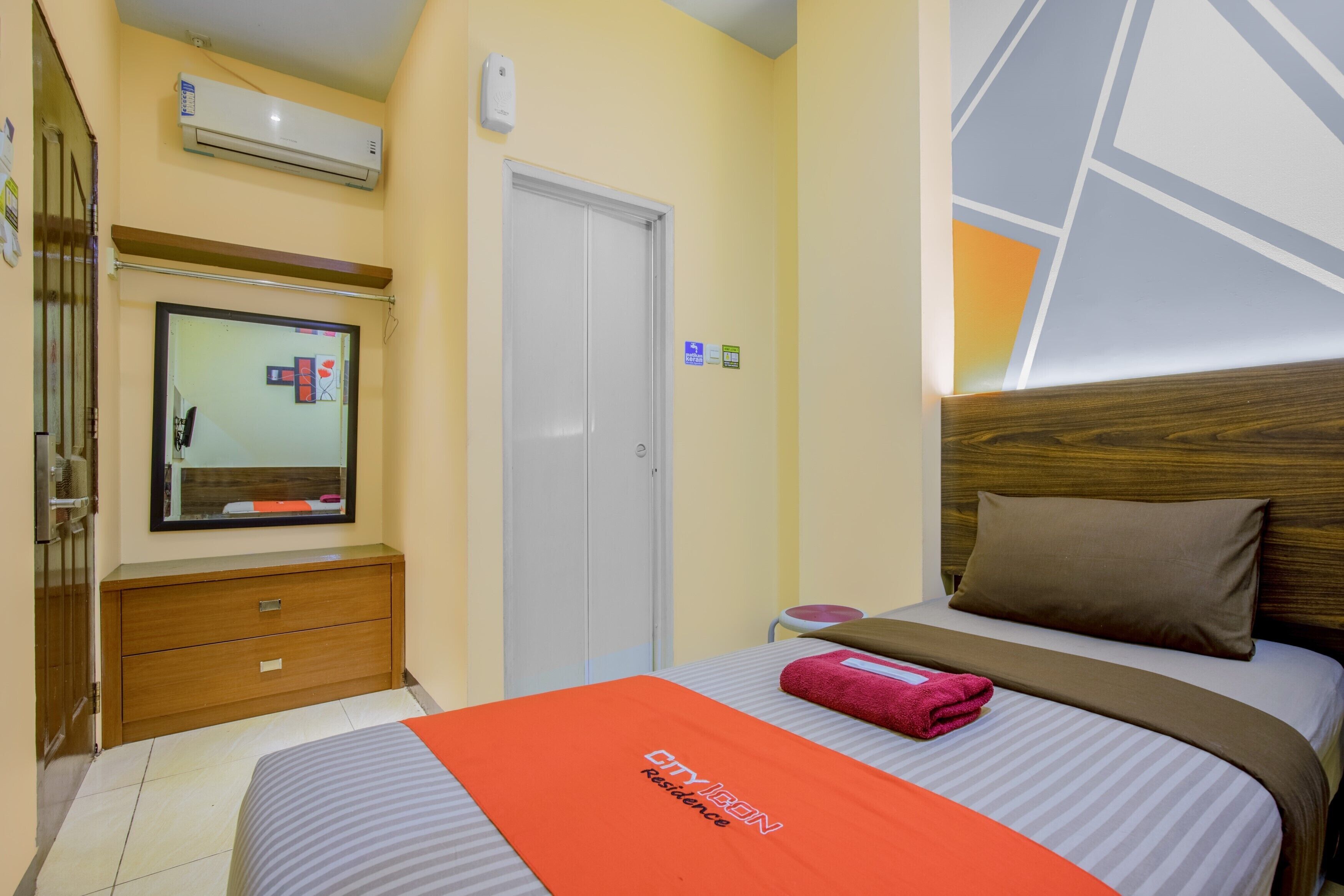 A hotel room with a bed, a wooden dresser, a mirror, an air conditioning unit, and a door.