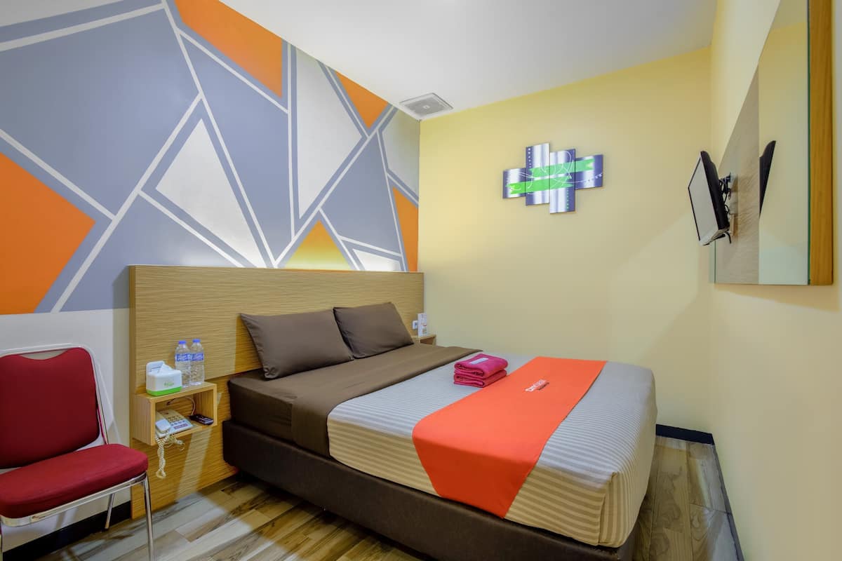 A hotel room with a bed, a red chair, a bedside table with a phone and water bottles, and a geometric wall design.