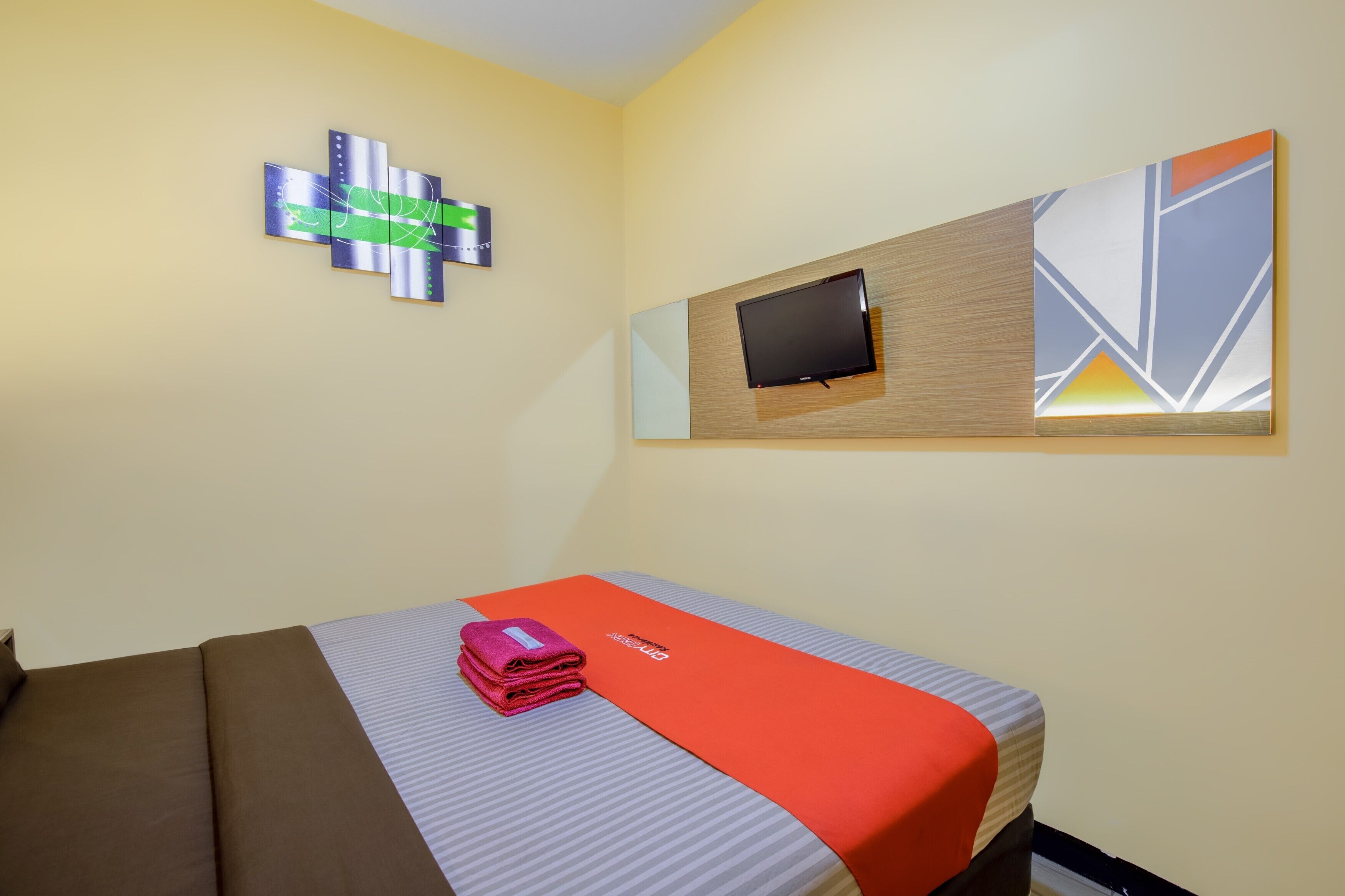 A hotel room with a bed, a flat-screen TV mounted on a wooden panel, and a wall art piece.
