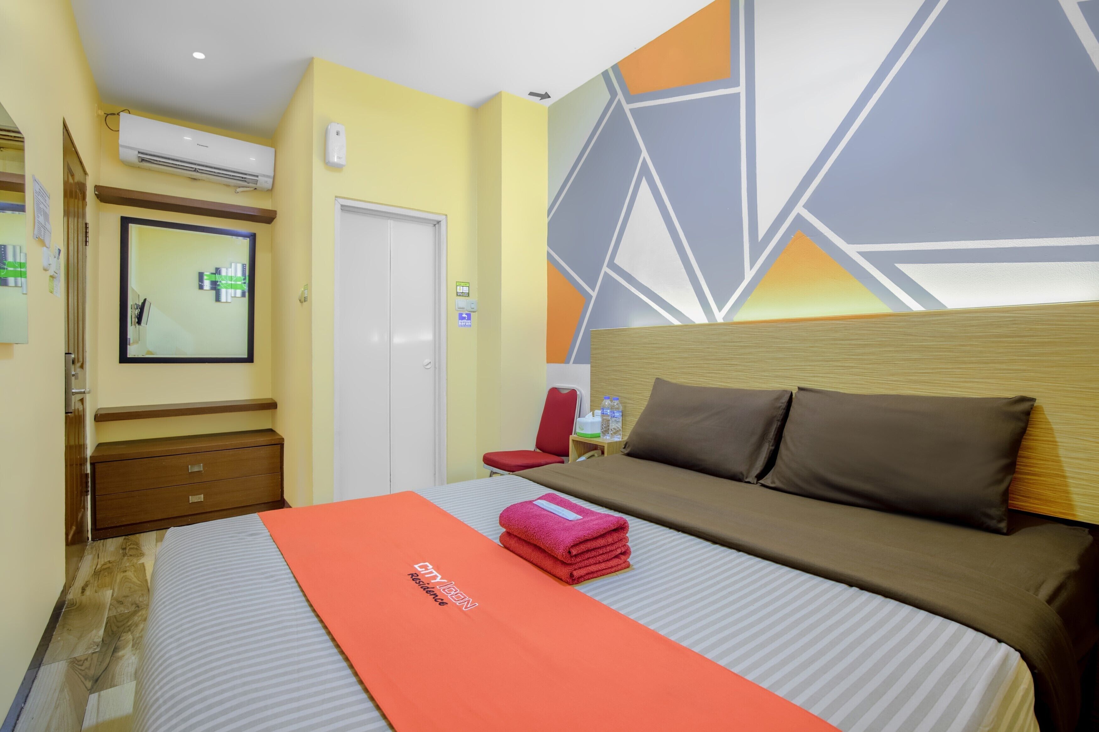 Una habitación de hotel con una cama, una silla roja, un cabecero de madera y un diseño geométrico en la pared.
