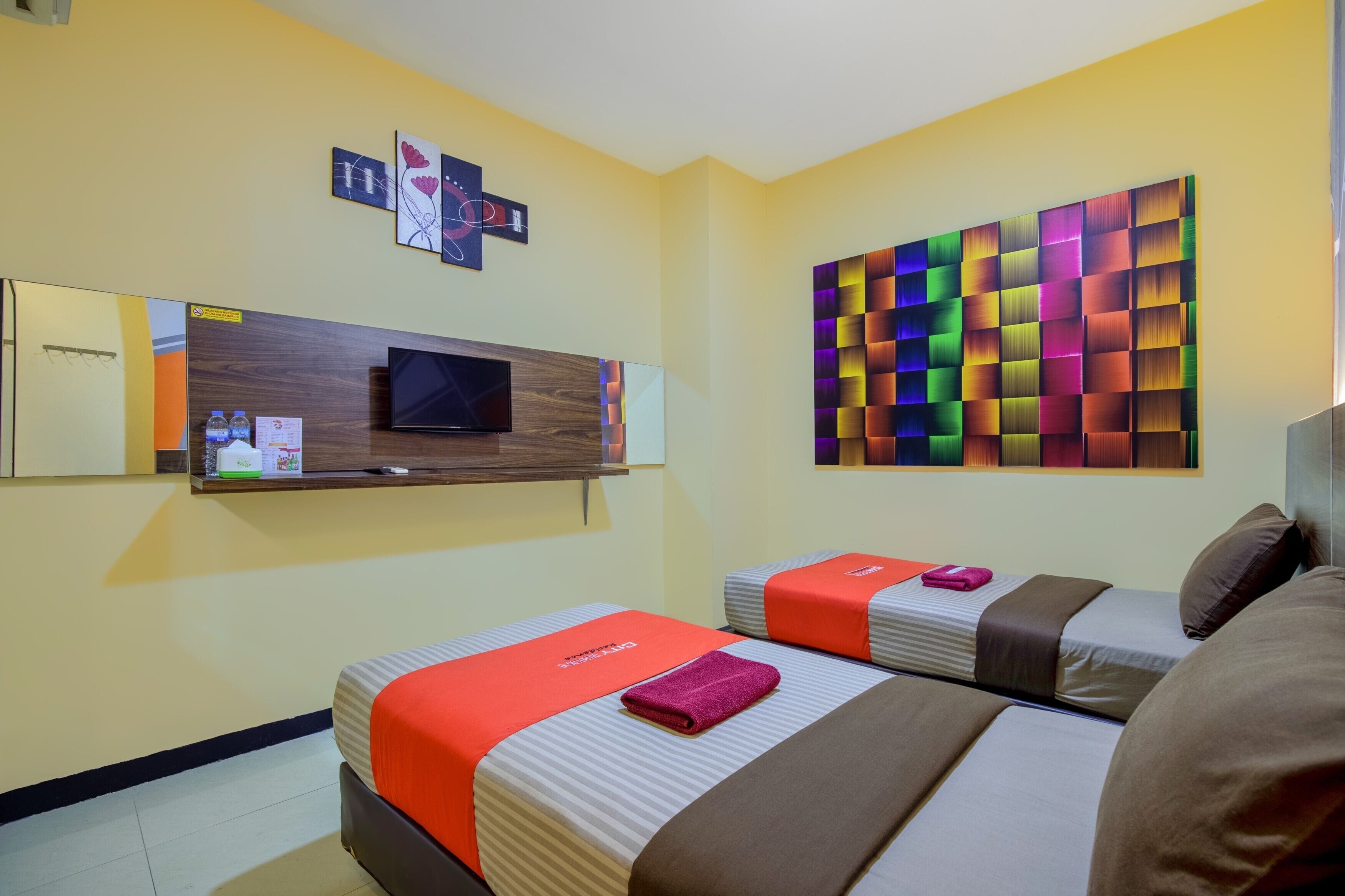 A hotel room with two beds, a TV, a colorful wall art, and a mounted shelf with water bottles and a sign.