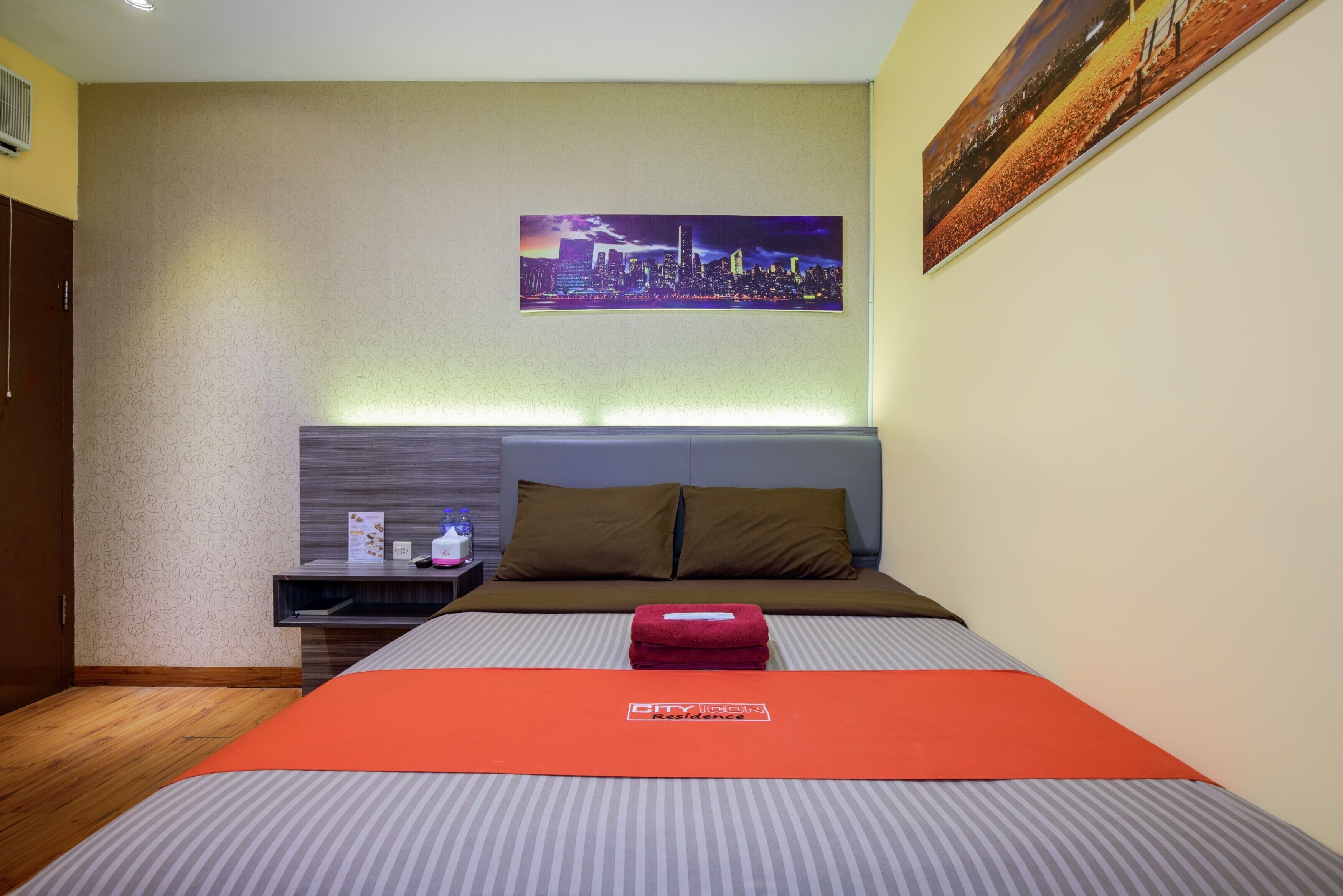 Habitació d'hotel amb un llit, una tauleta de nit, un quadre de paisatge urbà i una tovallola vermella sobre el llit.