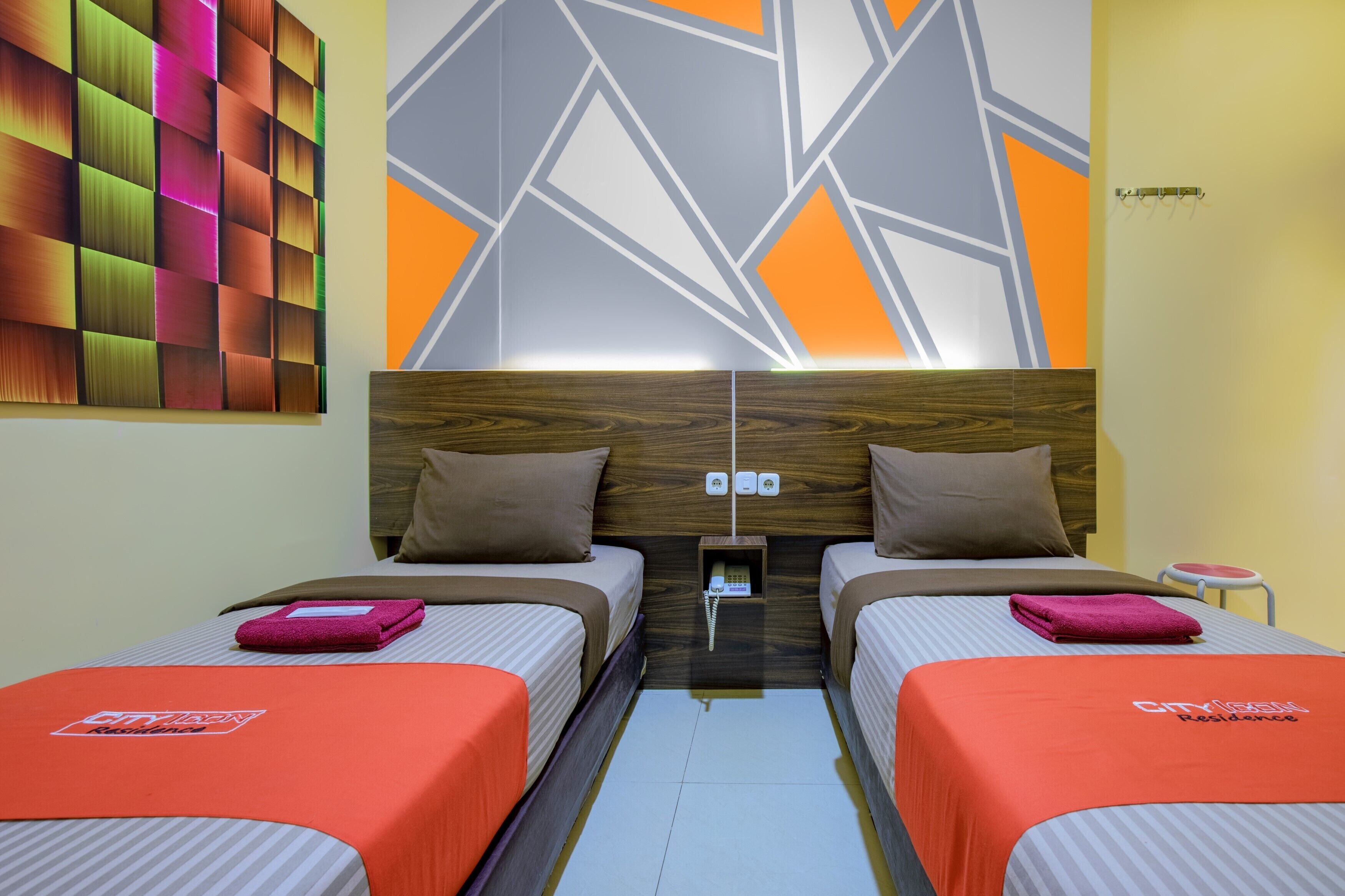 A hotel room with two beds, a wooden headboard, a colorful abstract wall, and a small table with a chair.