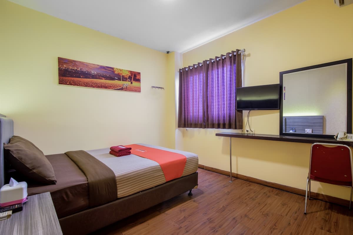 A hotel room with a bed, a desk with a computer, a TV, a window with curtains, and a painting on the wall.