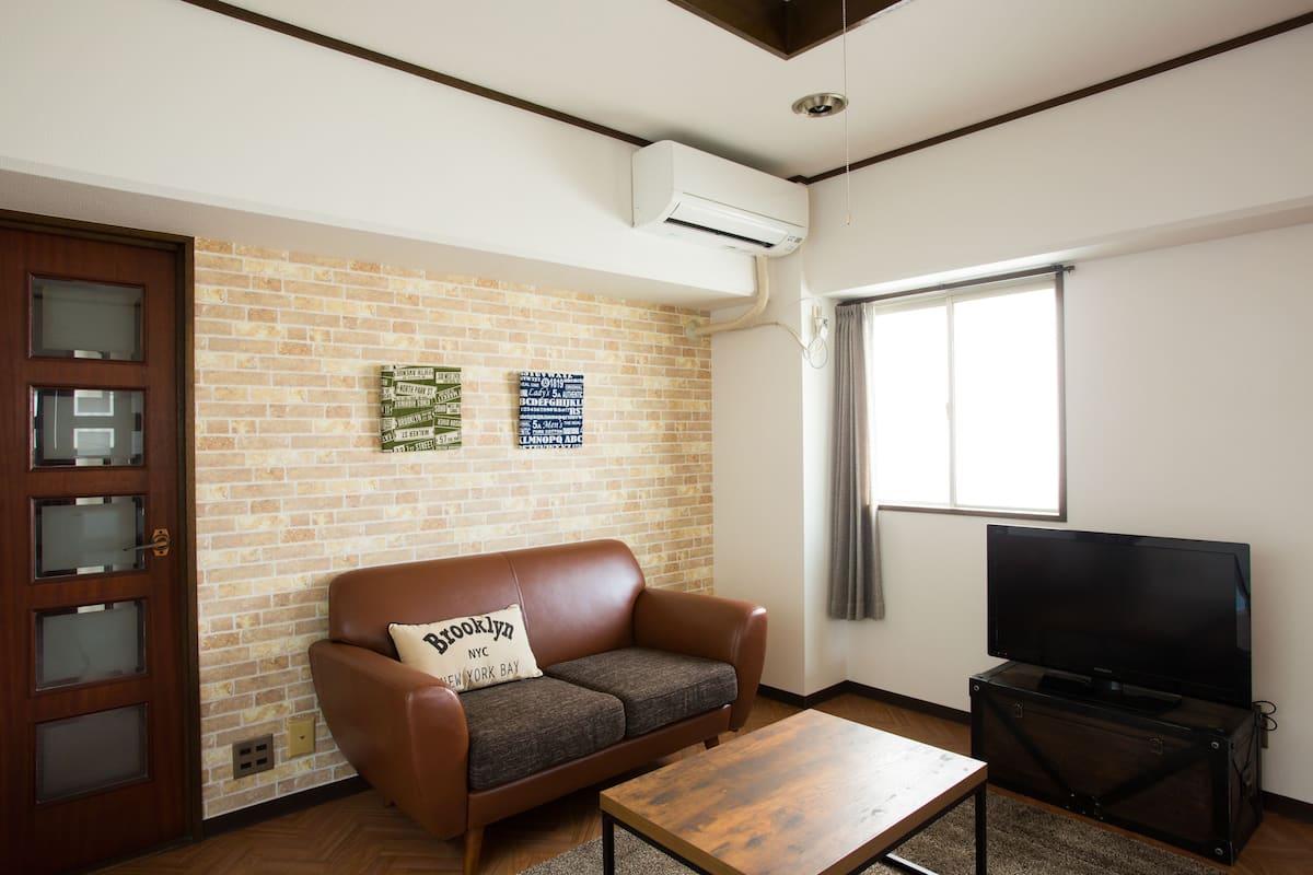 A living room with a brick wall, a brown leather sofa with a pillow, a wooden coffee table, a flat-screen TV on a stand, and a door with frosted glass panels.