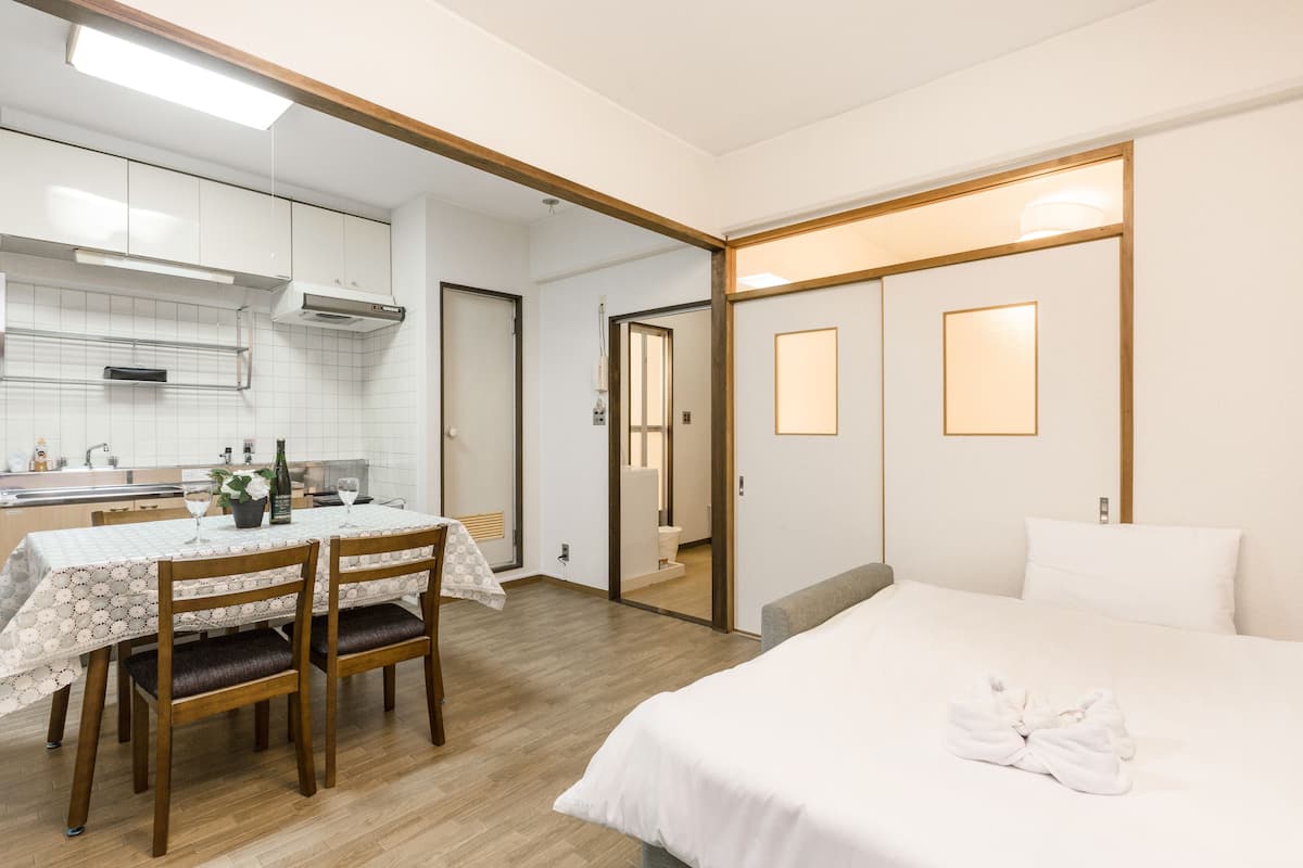 A hotel room with a bed, a dining table with chairs, a kitchen area, and a bathroom visible through an open door.