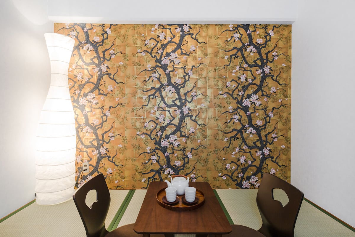 A traditional Japanese room with tatami flooring, a low table with a tray of white cups and a teapot, and a wall with a cherry blossom pattern.