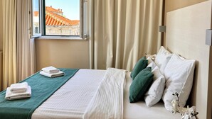 A hotel room with a bed, a window with curtains, a view of buildings, and a vase with flowers on the nightstand.