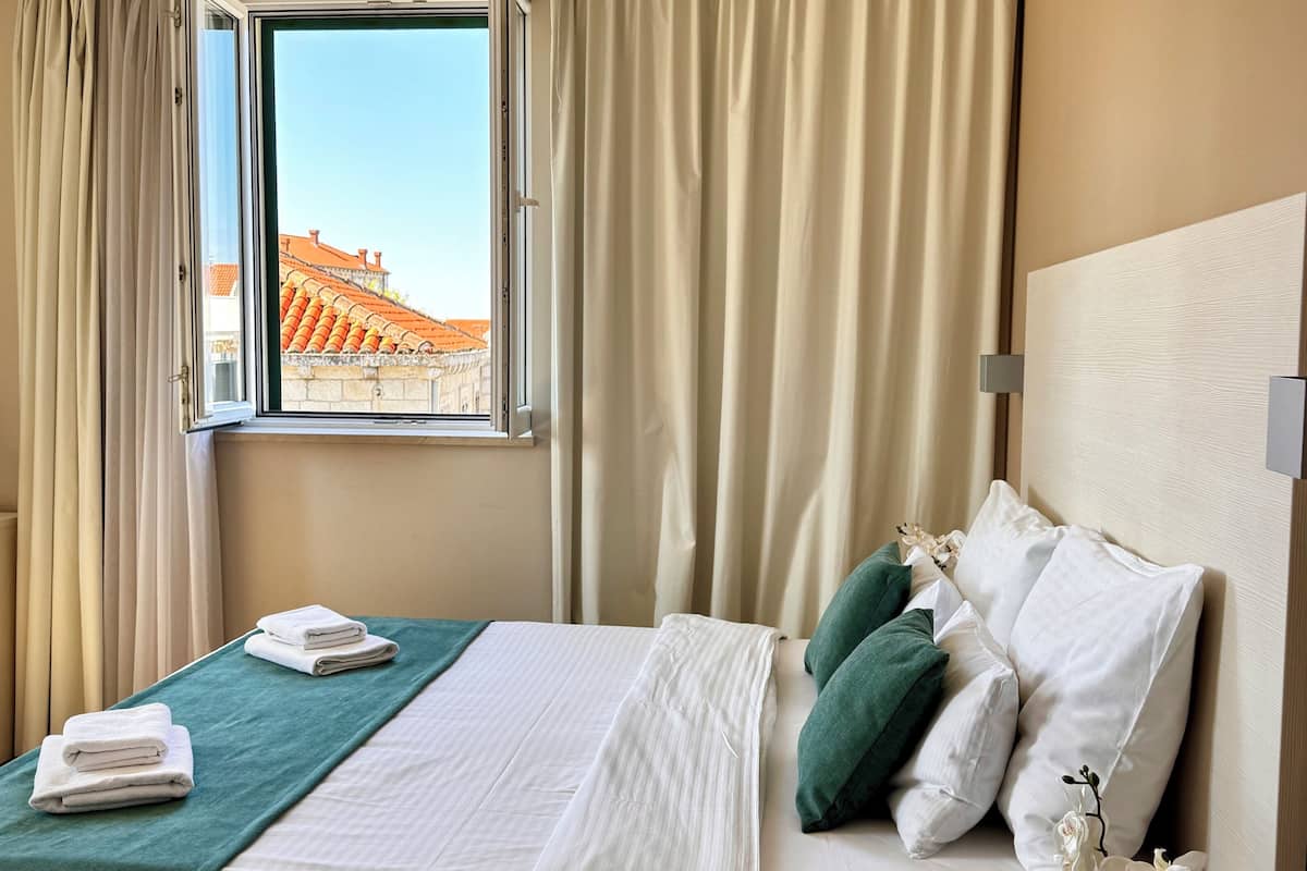 Una habitación de hotel con una cama, un ventanal con cortinas, vistas a edificios y un florero con flores en la mesita de noche.