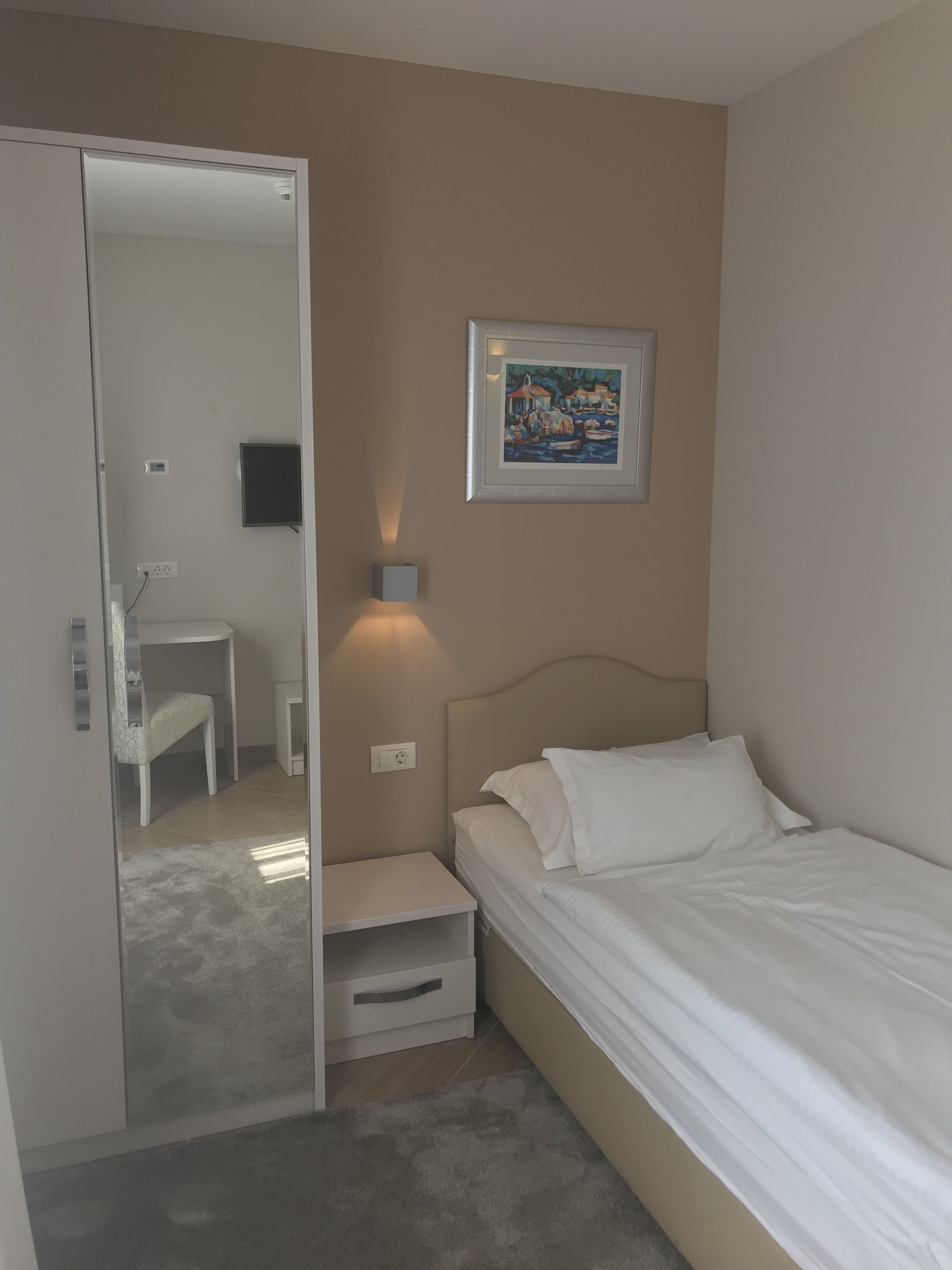 A bedroom with a bed, a bedside table, a nightstand, a chair, a desk, a painting, and a mirror.