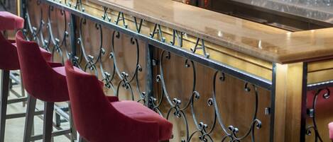 Zona de bar con mostrador de madera, taburetes rojos, lámparas colgantes y estantes con botellas y vasos.