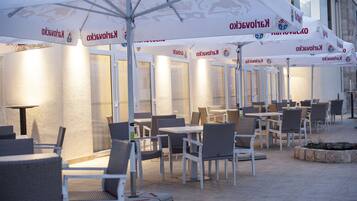 An outdoor seating area with white tables and chairs, umbrellas with logos, and a stone wall in the background.