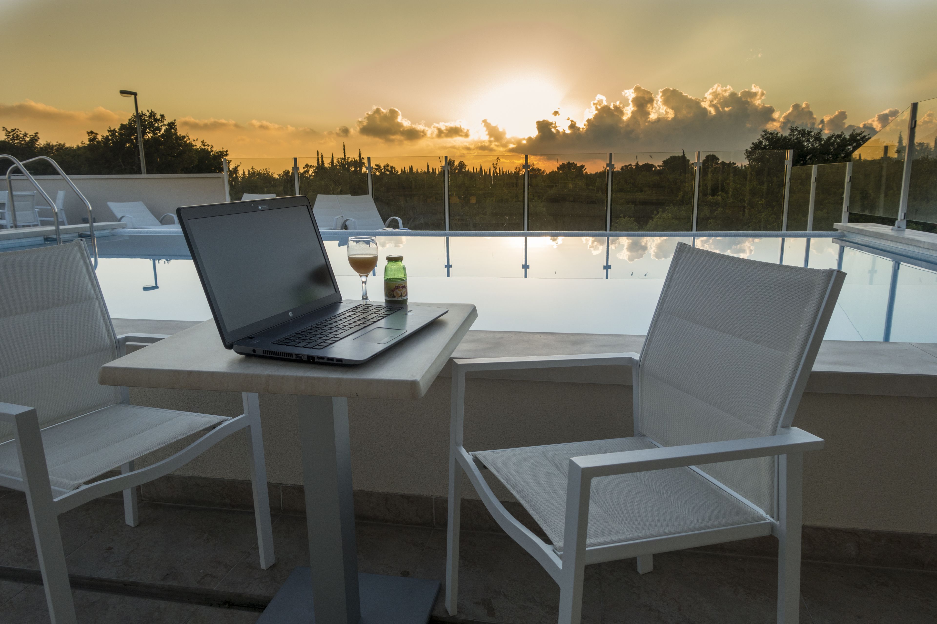 Ein Laptop auf einem Tisch, ein Glas, zwei weiße Stühle und Blick auf einen Sonnenuntergang über dem Wasser.
