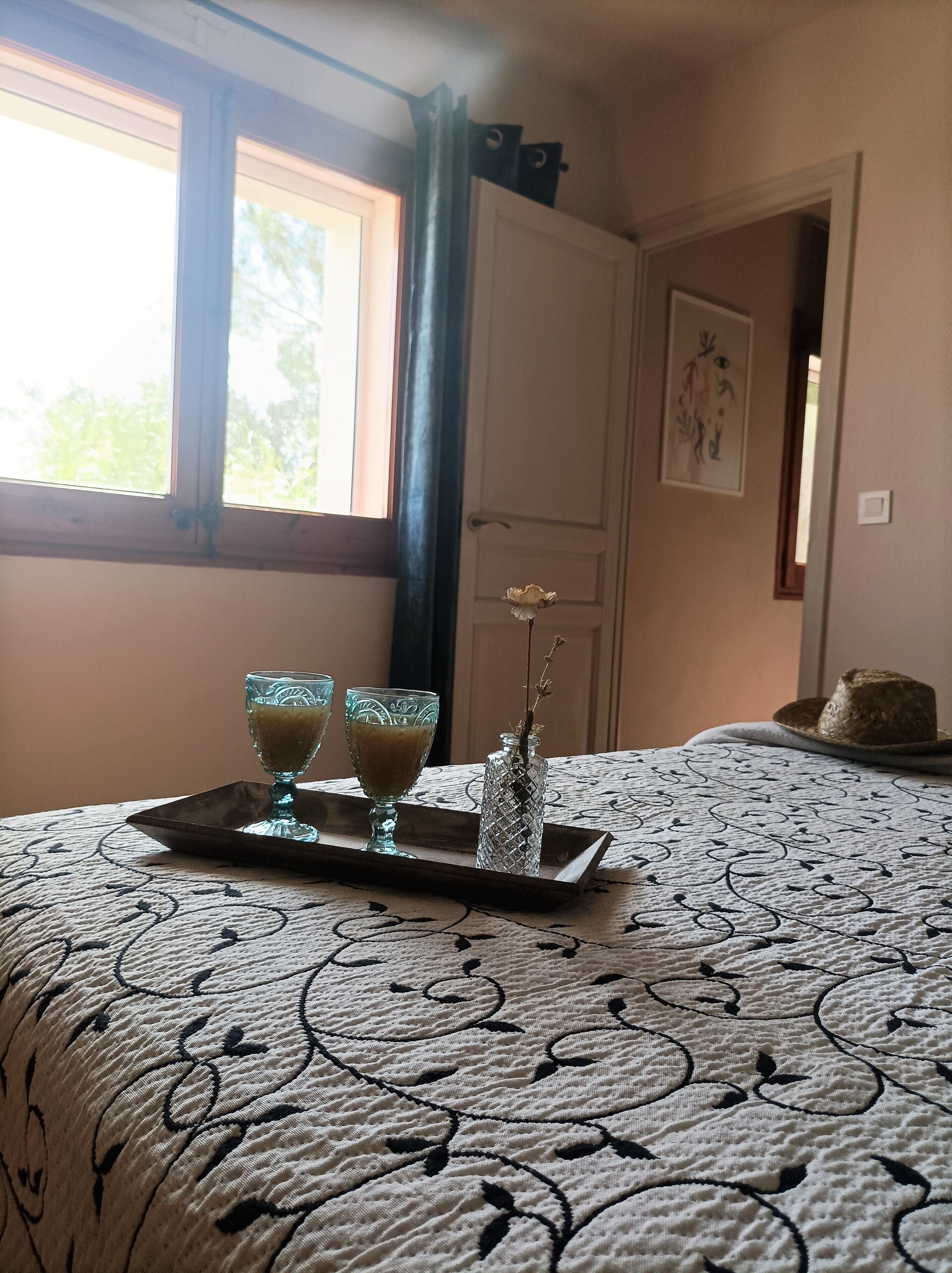 Una cama bien tendida con una colcha estampada, una bandeja con dos vasos de una bebida y un florero con una rama.
