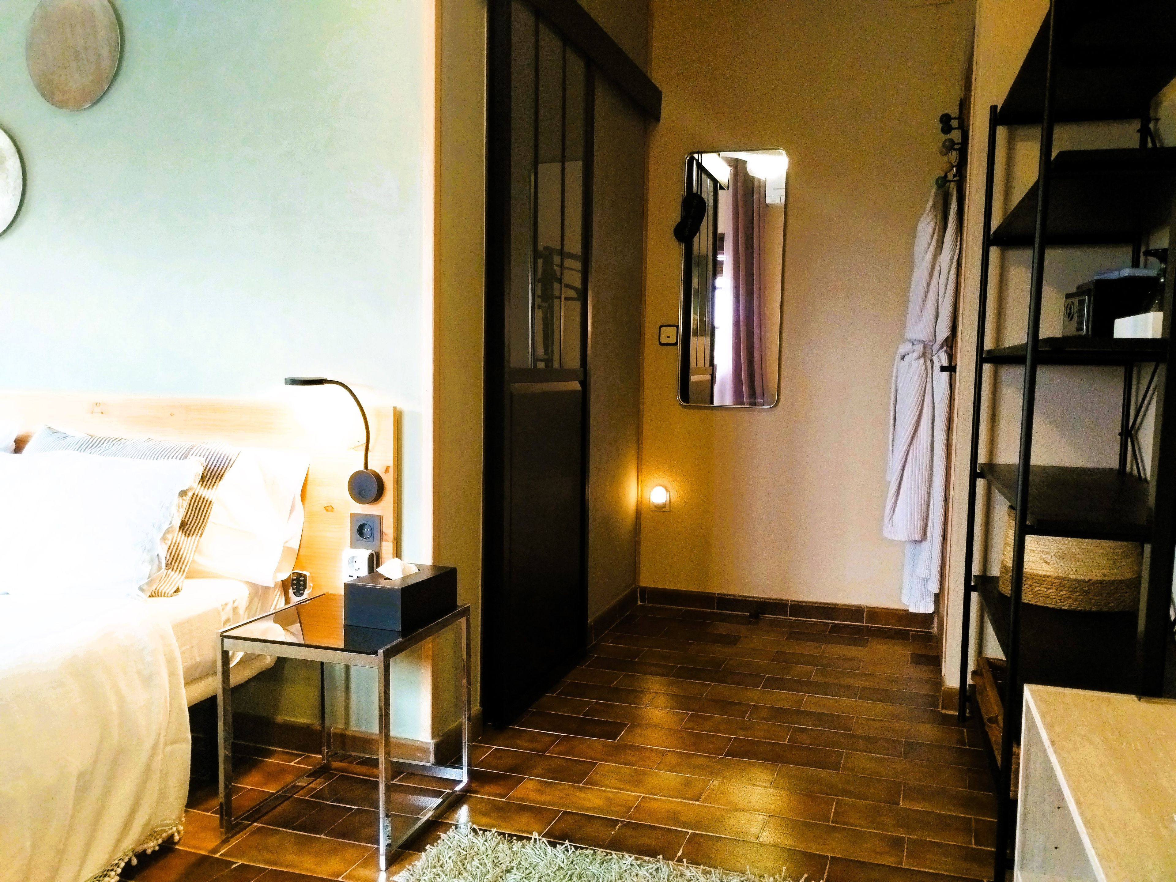 Una habitación de hotel con cama, mesita de noche, lámpara, espejo y un pequeño estante.