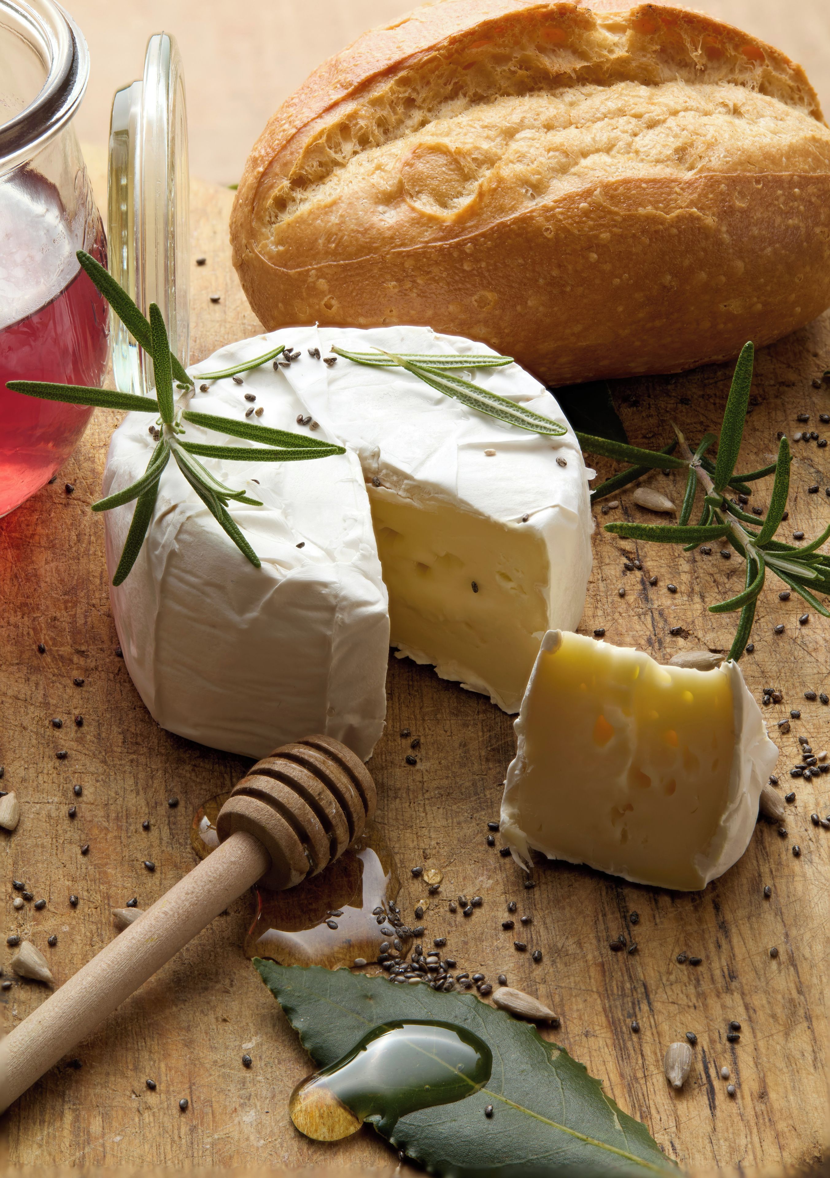 Un queso redondo, una hogaza de pan y un cucharón de miel sobre una superficie de madera con semillas y hierbas esparcidas.