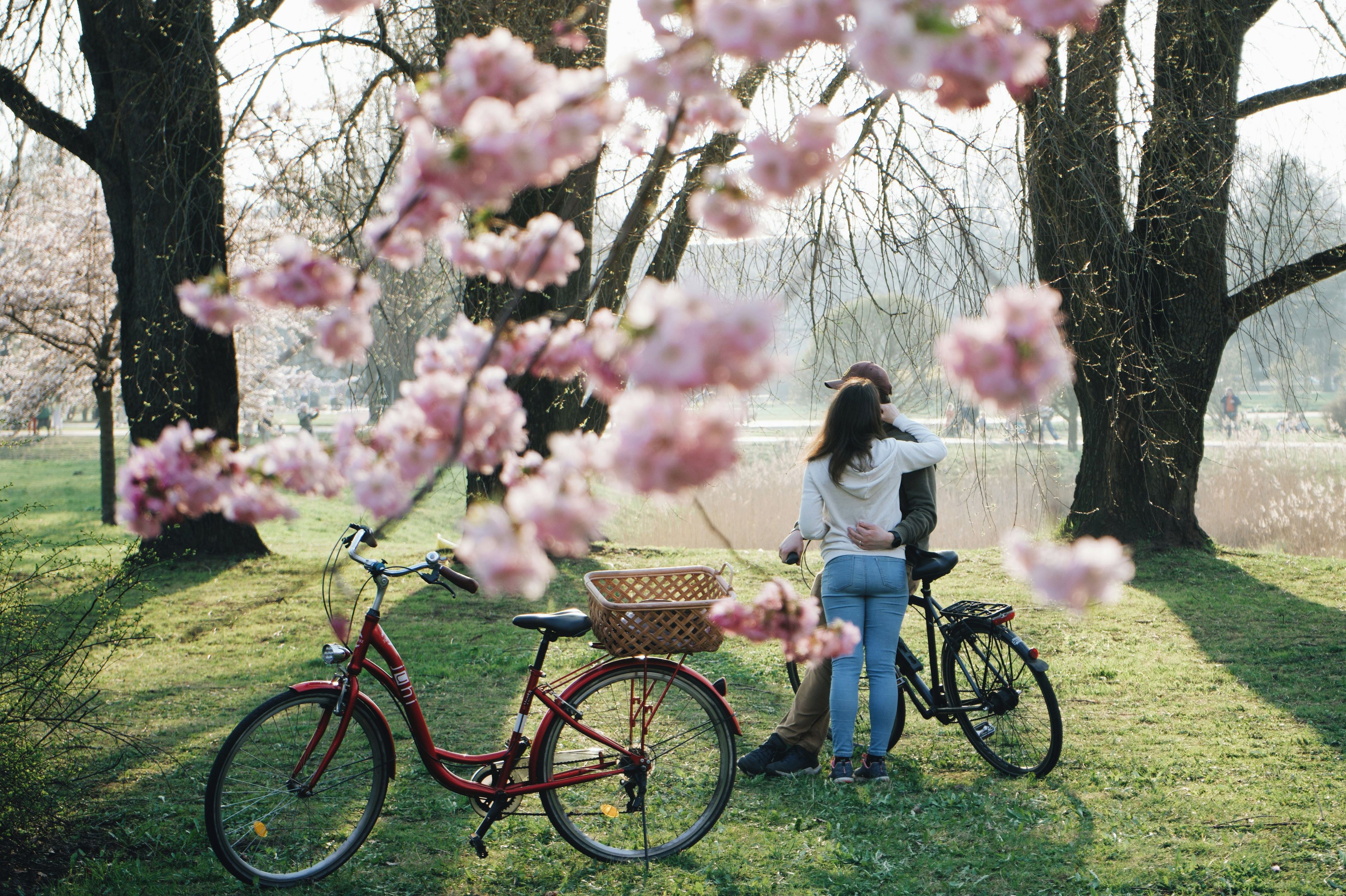 Un vélo rouge avec un panier, une personne debout à côté, et des fleurs de cerisier au premier plan.
