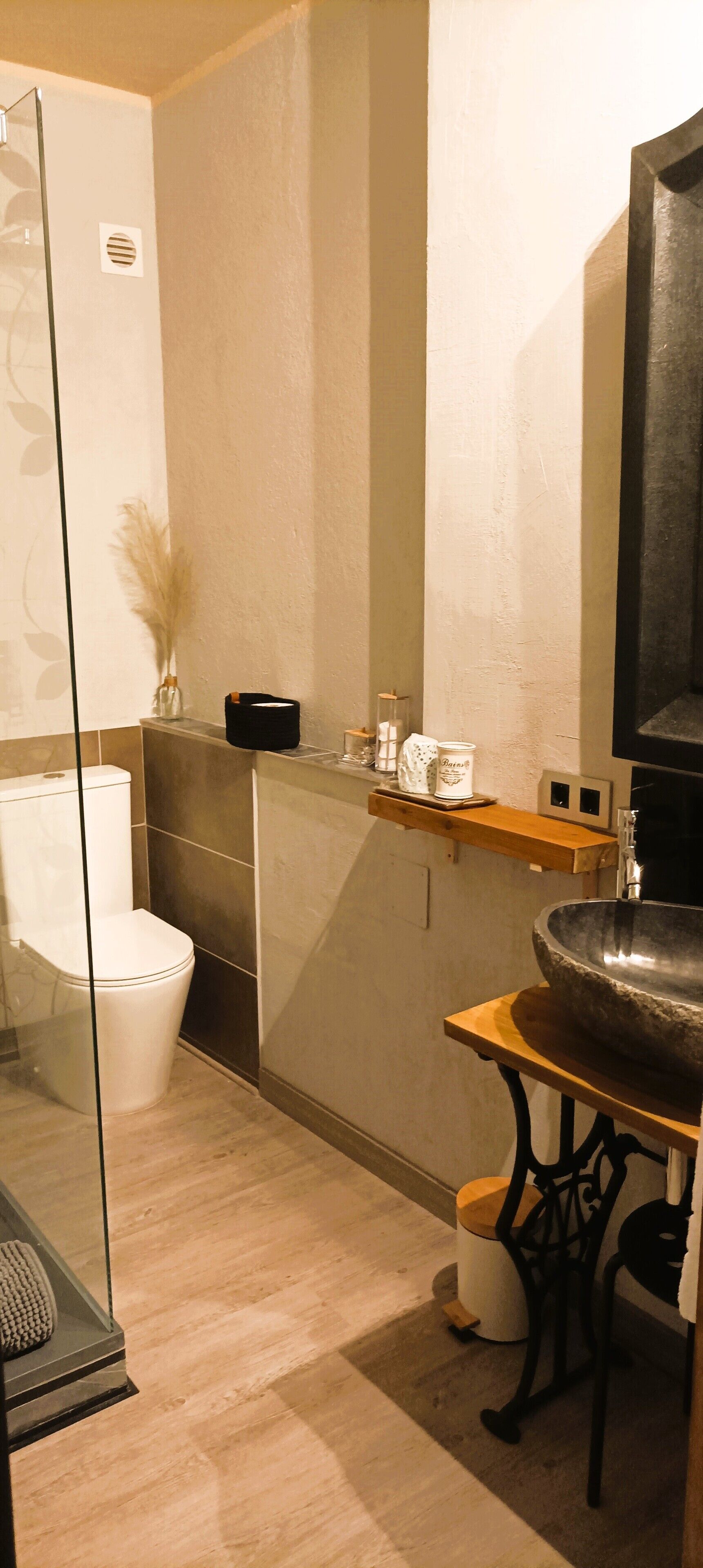 A modern bathroom with a glass shower enclosure, a white toilet, a large stone sink, and a wooden shelf with toiletries.