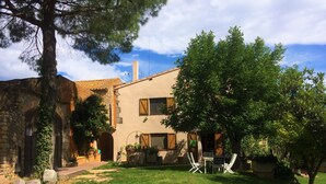 A two-story house with a stone wall on the left, a tiled roof, and a grassy yard with a tree and outdoor furniture. - La Masia Costa Brava (Garrigas)