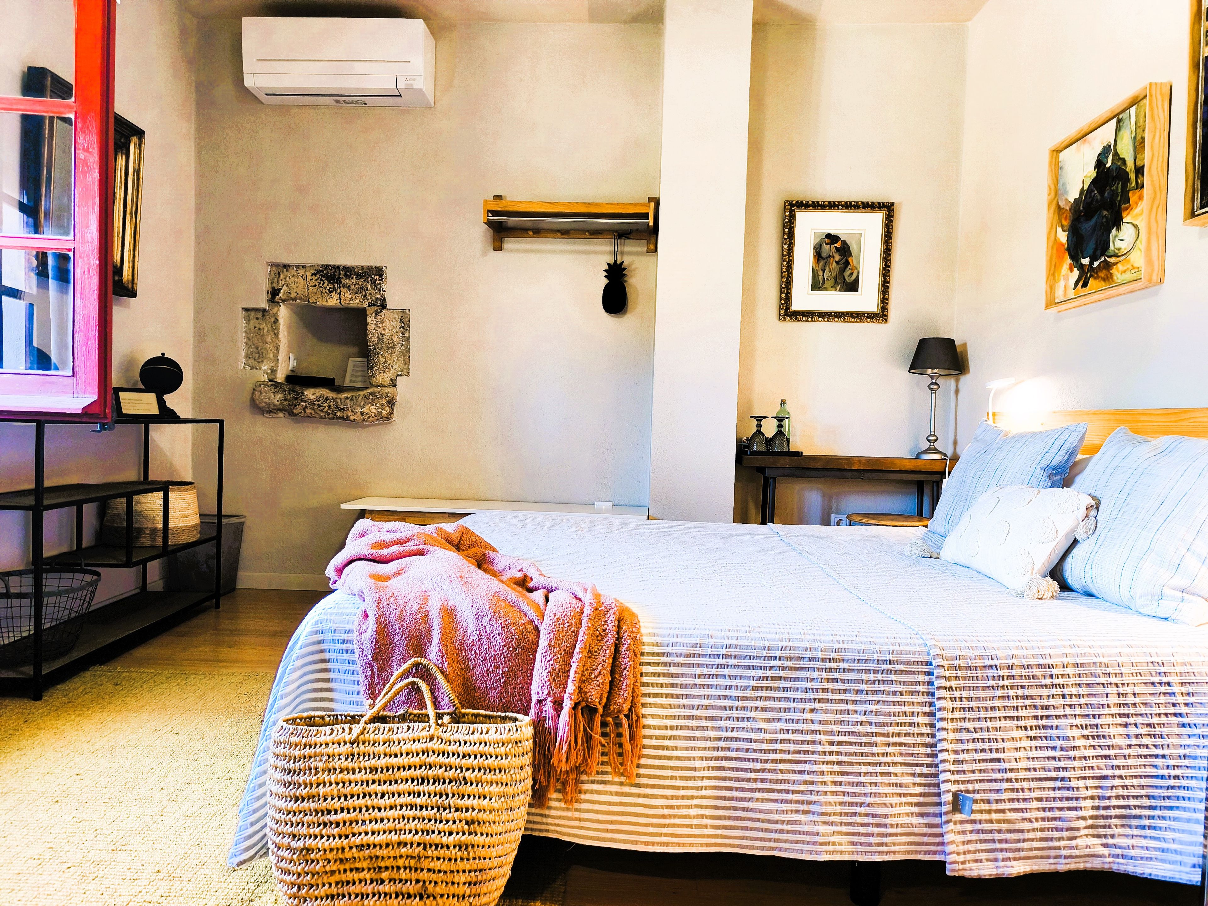 Un dormitorio con cama, mesita de noche, lámpara, aire acondicionado instalado en la pared y un cuadro enmarcado.