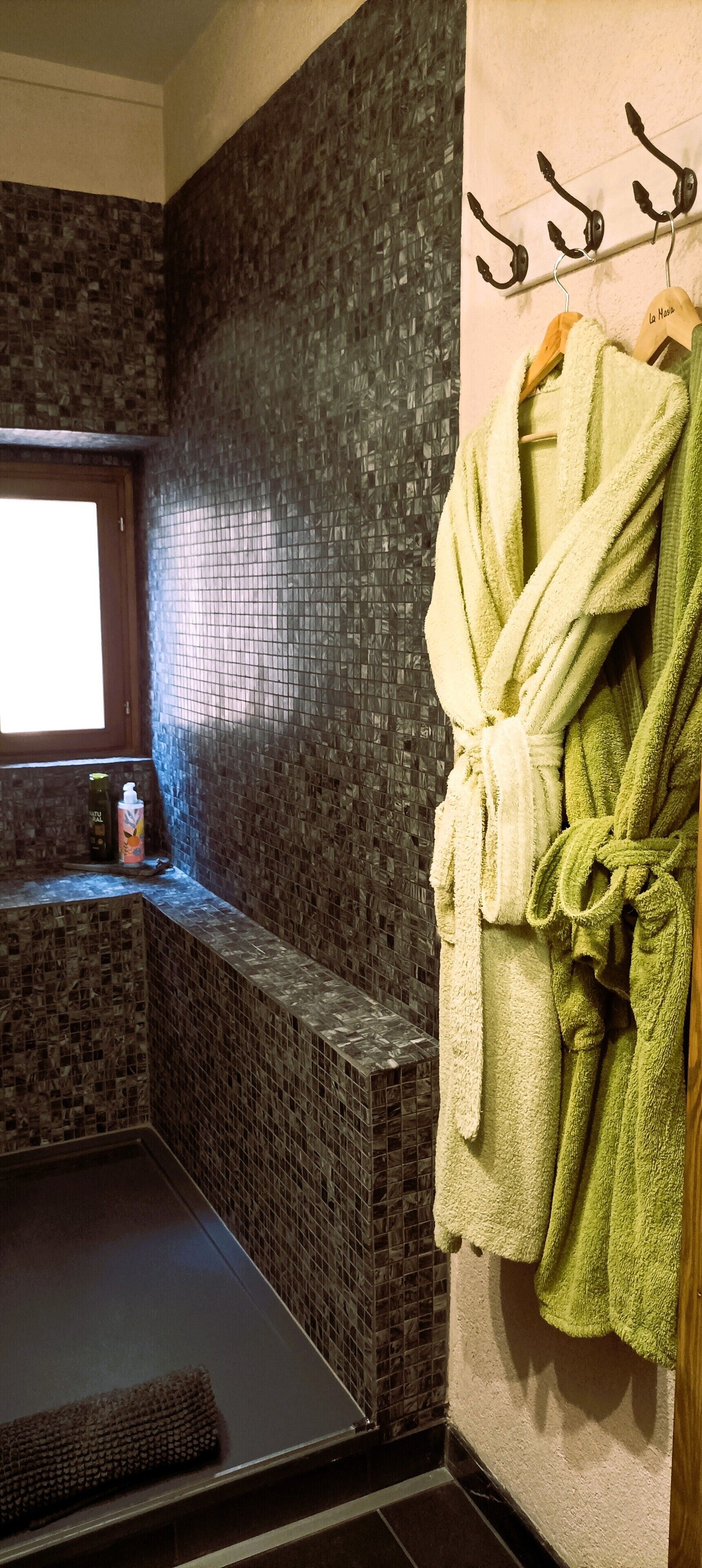 Un baño con paredes de mosaico, una ventana y un perchero con batas.