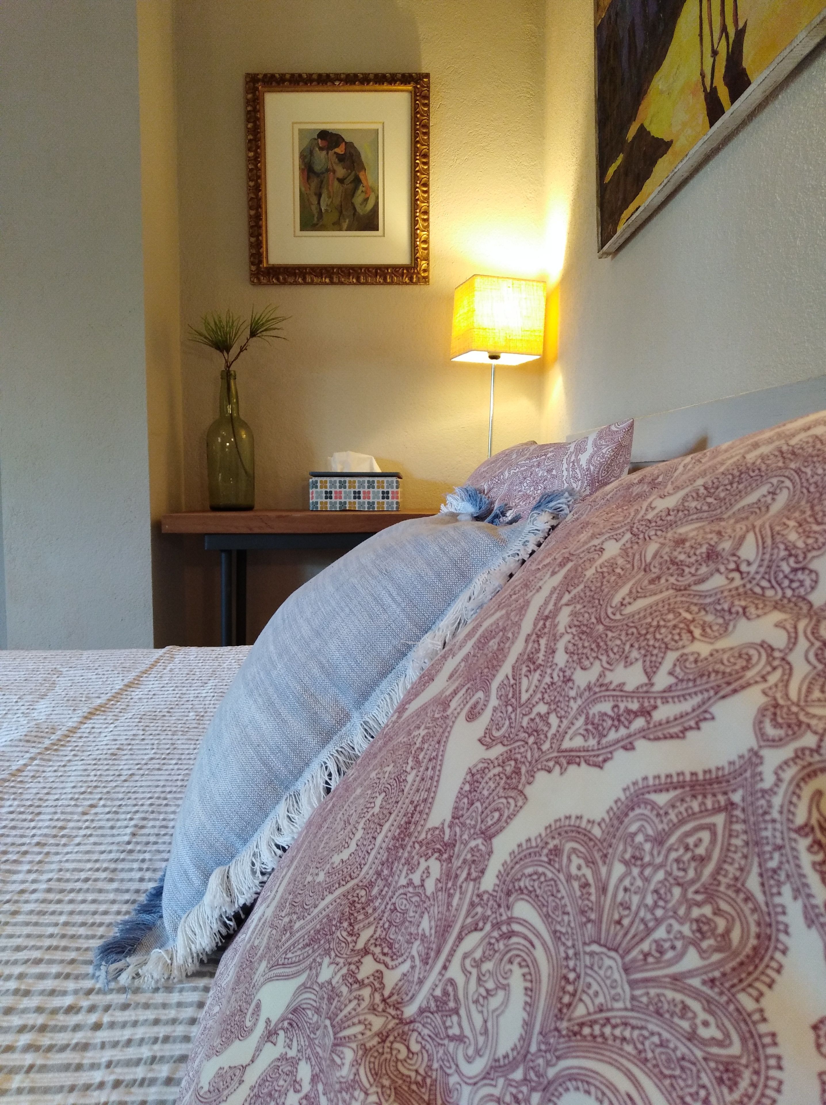 Una cama bien hecha con ropa de cama estampada, una lámpara de noche, un cuadro en la pared y una planta pequeña en una mesita de noche.