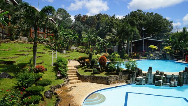 A pool surrounded by a landscaped garden with stone steps leading to the pool, surrounded by tropical vegetation and a building in the background. - Bakasyunan Resort Tanay (Tanay)